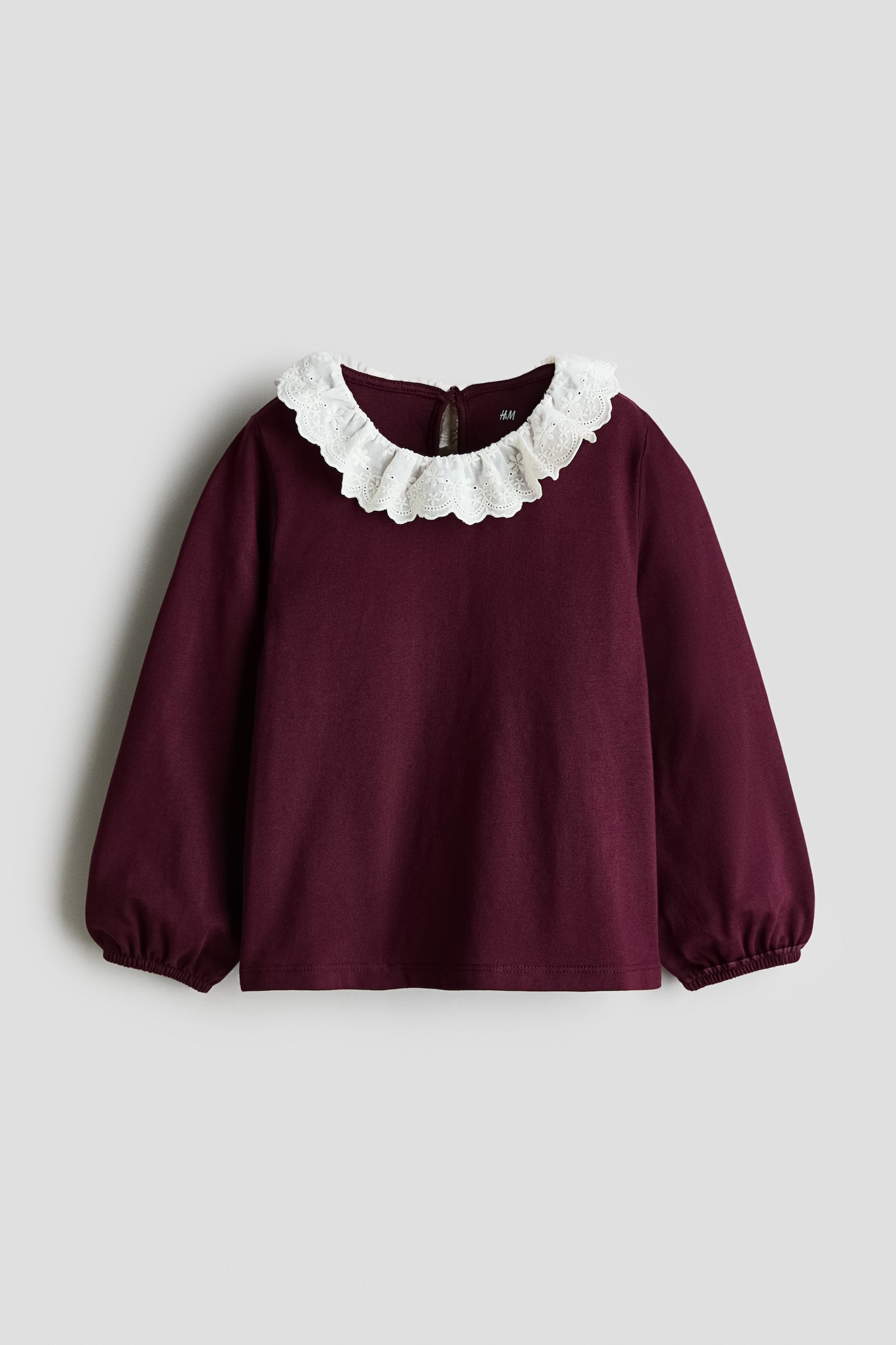 Un top bordeaux a maniche lunghe Ã¨ mostrato frontalmente, con un colletto bianco in pizzo sangallo arricciato con bordi smerlati. Il top presenta maniche lunghe con polsini elasticizzati arricciati e una piccola apertura a goccia sul retro del collo.