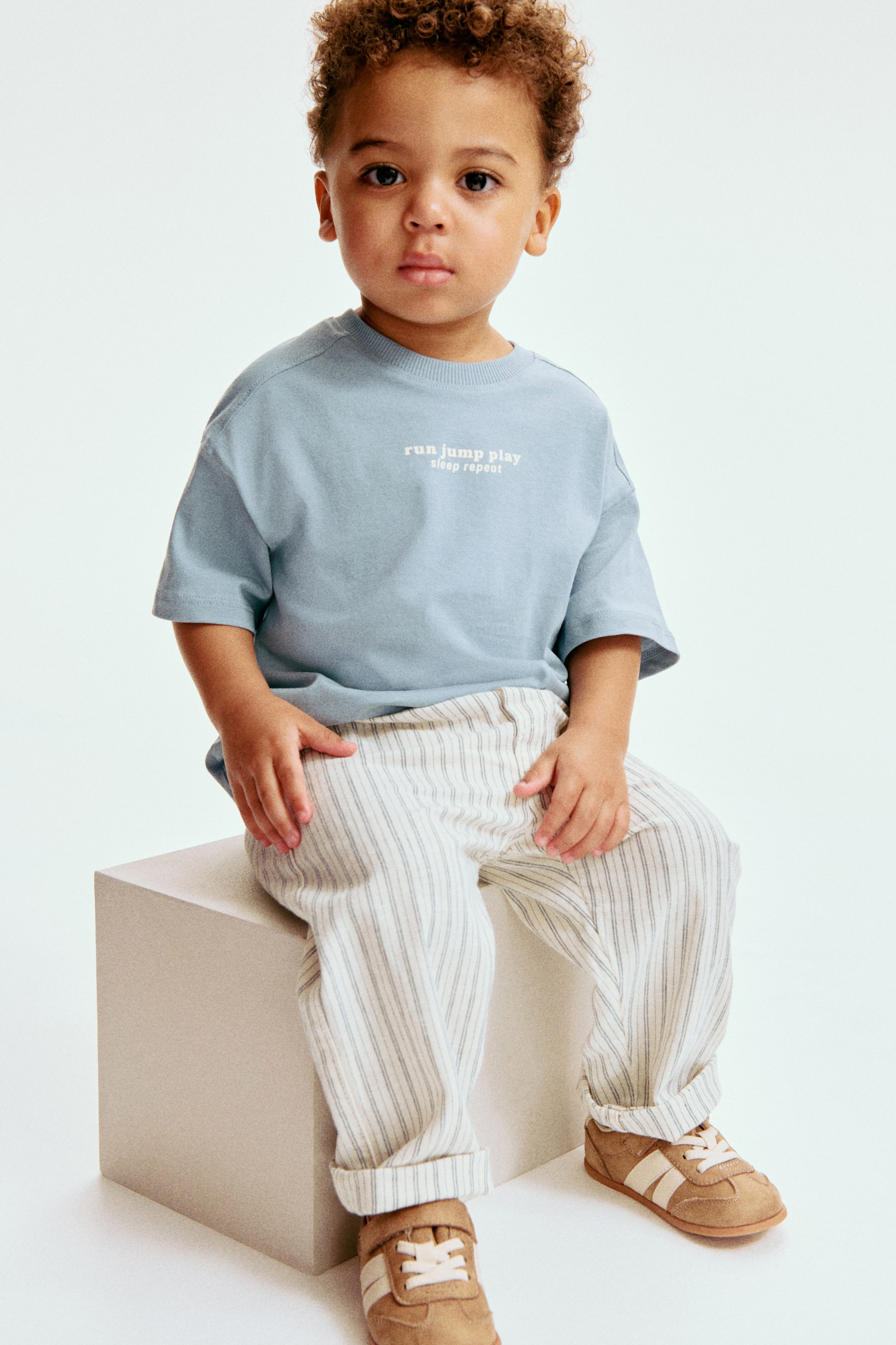 Grösseres Bild ansehen: Ein hellblaues T-Shirt mit geripptem Rundhalsausschnitt und weißem Text auf der Brust, der "run jump play sleep repeat" lautet. Dazu passen hellfarbene, vertikal gestreifte Hosen, die locker sitzen und an den Knöcheln umgeschlagen sind, sowie beige Sneakers mit weißen Seitenstreifen und Klettverschlüssen.