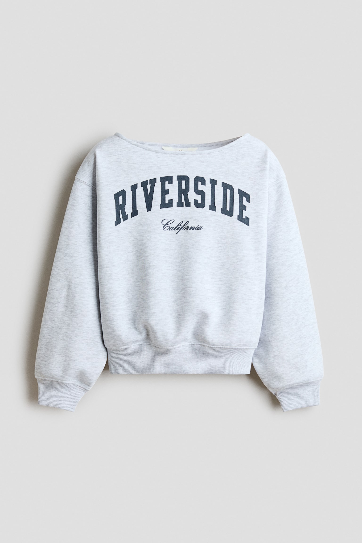 En ljusgrÃ¥ melerad sweatshirt visas framifrÃ¥n, med en bred bÃ¥thalsringning, nedsÃ¤nkta axlar och ribbade muddar vid Ã¤rmsluten och nederkanten. PÃ¥ brÃ¶stet visas ordet âRIVERSIDEâ i bÃ¥gformade marinblÃ¥ blockbokstÃ¤ver med âCaliforniaâ tryckt i kursiv stil nedanfÃ¶r.