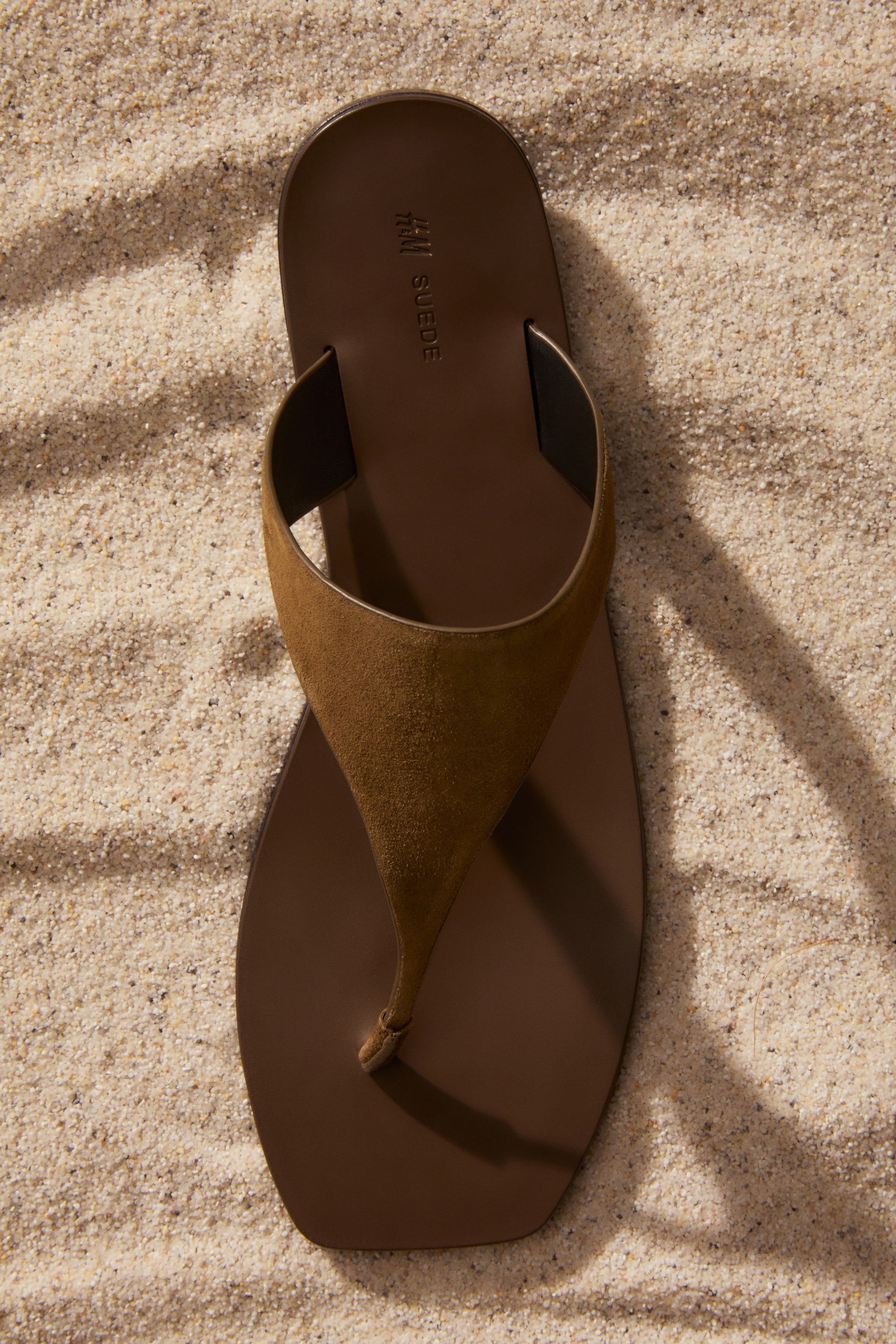 Grotere afbeelding bekijken: Een bruine sandaal ligt plat, gezien vanuit een bovenaanzicht. Het gladde voetbed toont 'H&M SUEDE' in reliëf nabij de bovenkant, en een brede, tan suède V-vormige band vormt het teenslipperontwerp.