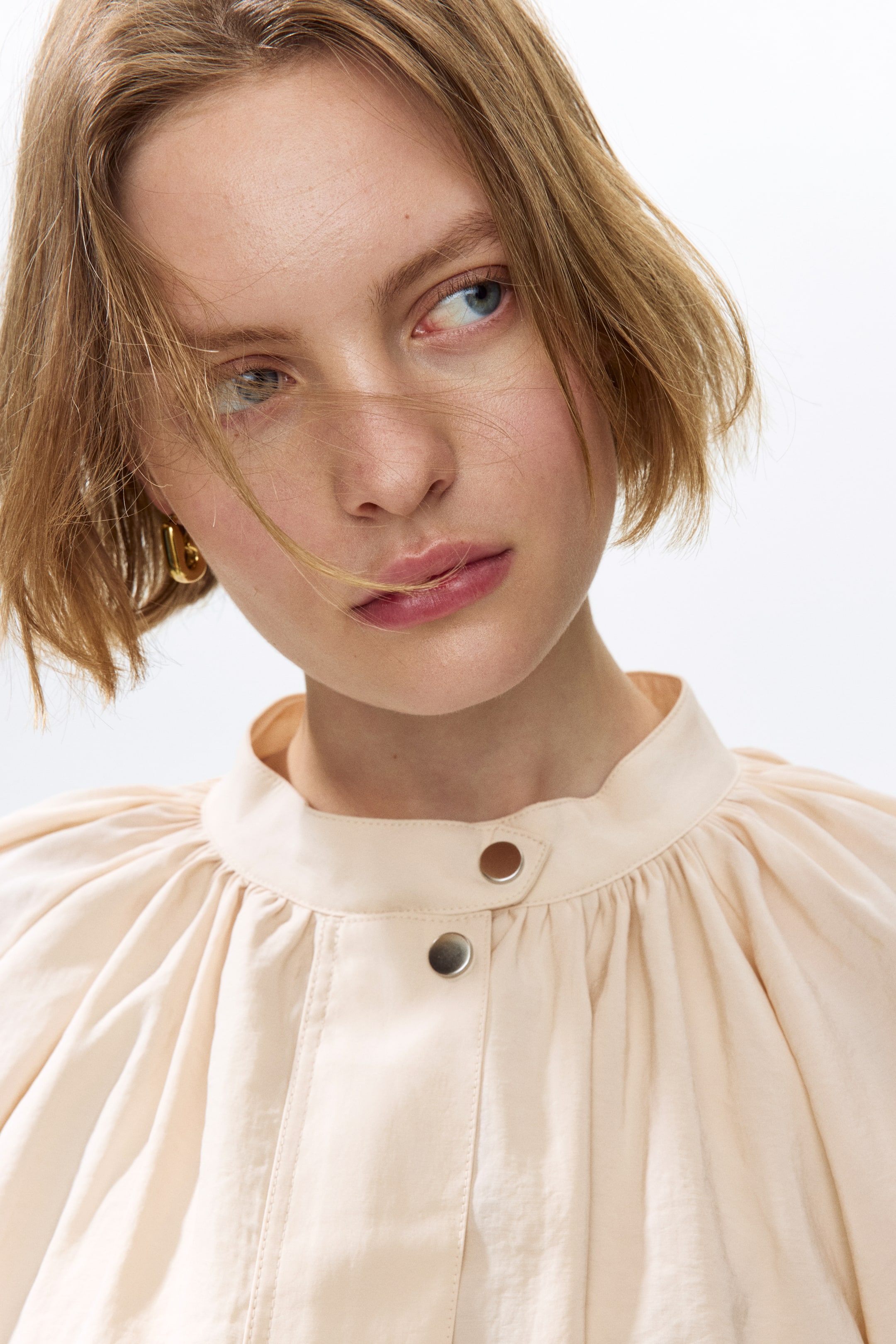 View larger image: The model is donning an oversized jacket crafted from a pale peach modal weave. It showcases a band collar with gathered detailing around the neckline and a front wind flap secured with two visible press-studs.