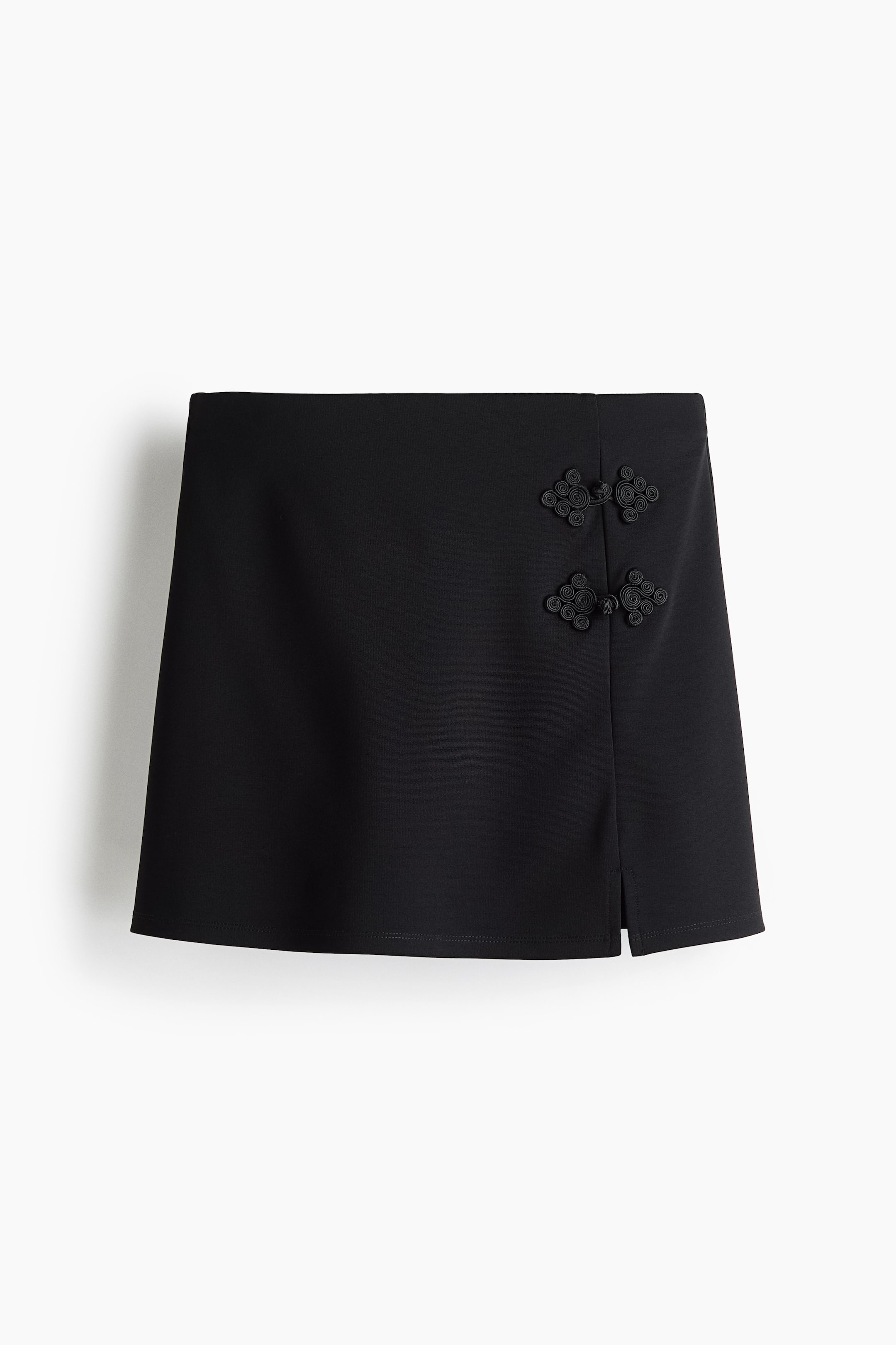 Grotere afbeelding bekijken: Dit korte rokje van viscosemix in zwart is van voren te zien en heeft een rechte silhouet met een platte tailleband. Twee decoratieve zwarte frog-sluitingen accentueren de faux-wrapnaad boven een kleine zijsplit aan de zoom.