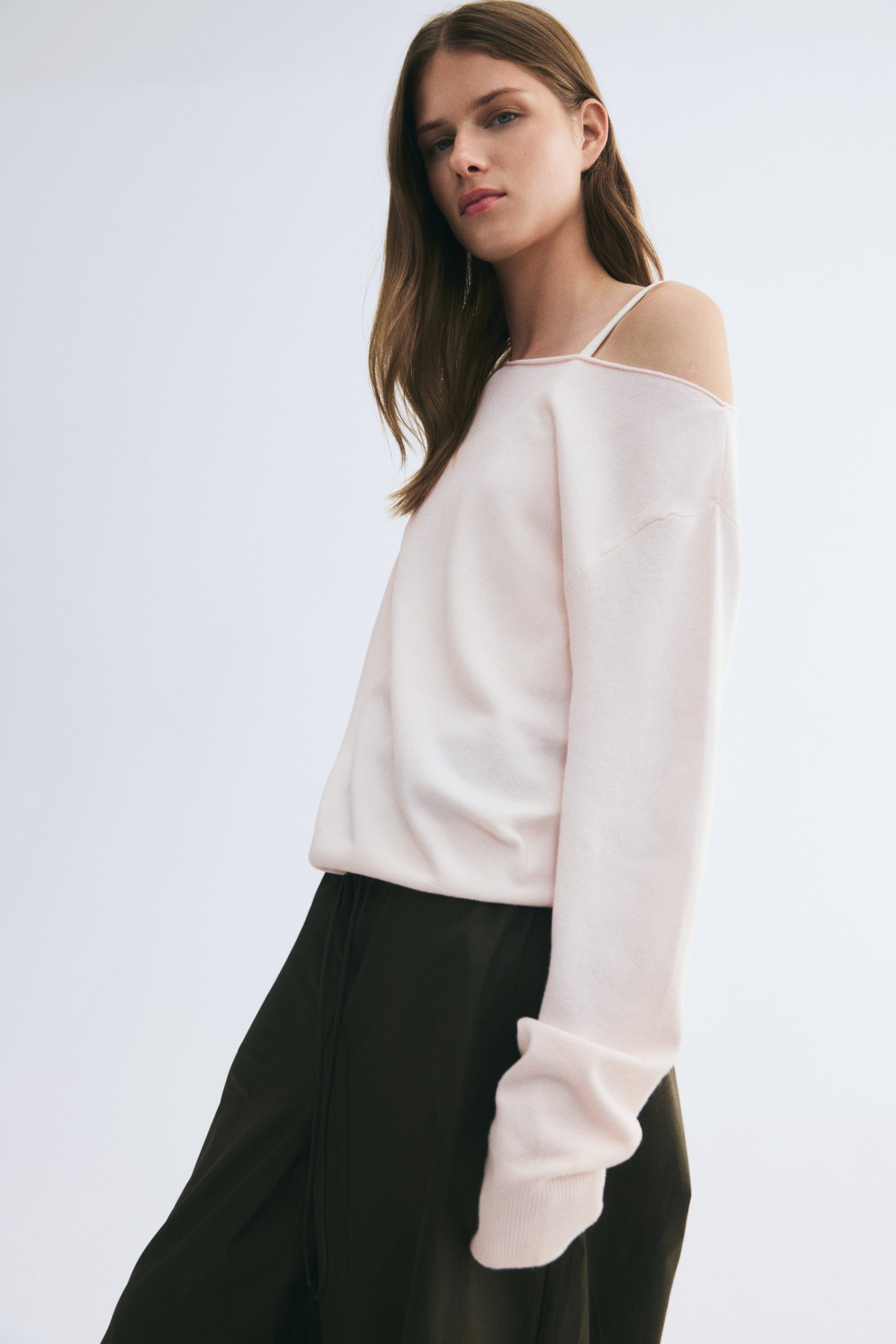 View larger image: An off-the-shoulder knit sweater in a light pink shade is being worn by the model, paired with dark green trousers. The sweater features a roll-edge neckline exposing one shoulder, dropped shoulders, and long sleeves with ribbed cuffs.