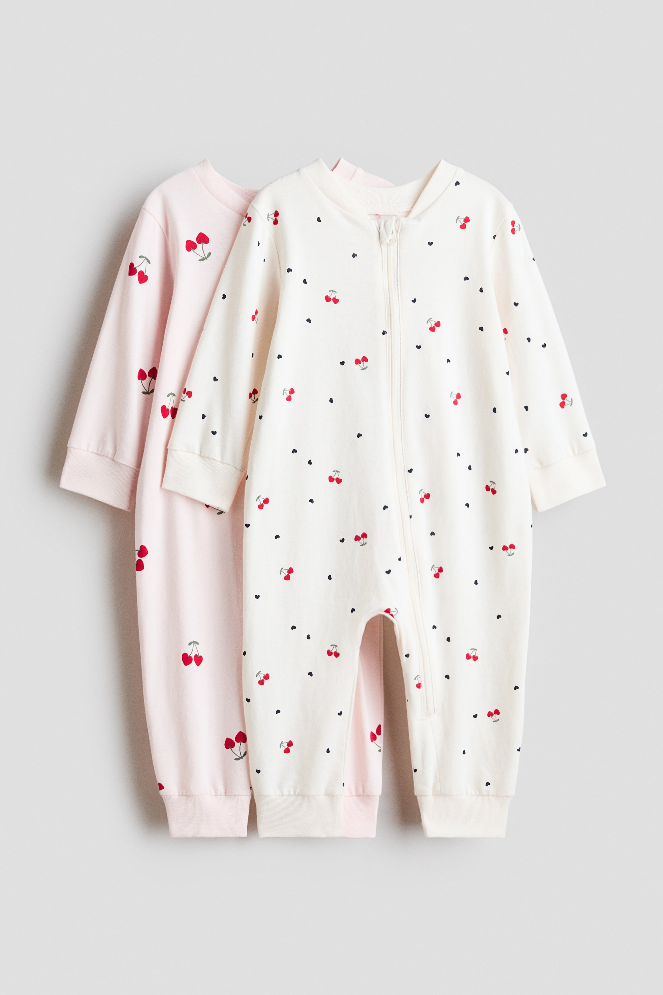 Ver imagem maior: Dois babygrows estão deitados planos, virados para a frente, com um ligeiramente sobrepondo o outro. O babygrow da frente é de cor creme, apresentando um padrão de cerejas vermelhas e pequenos corações pretos, um fecho de correr de comprimento total e punhos canelados nos pulsos e tornozelos, enquanto o babygrow rosa atrás exibe um padrão repetido de cerejas com punhos canelados.