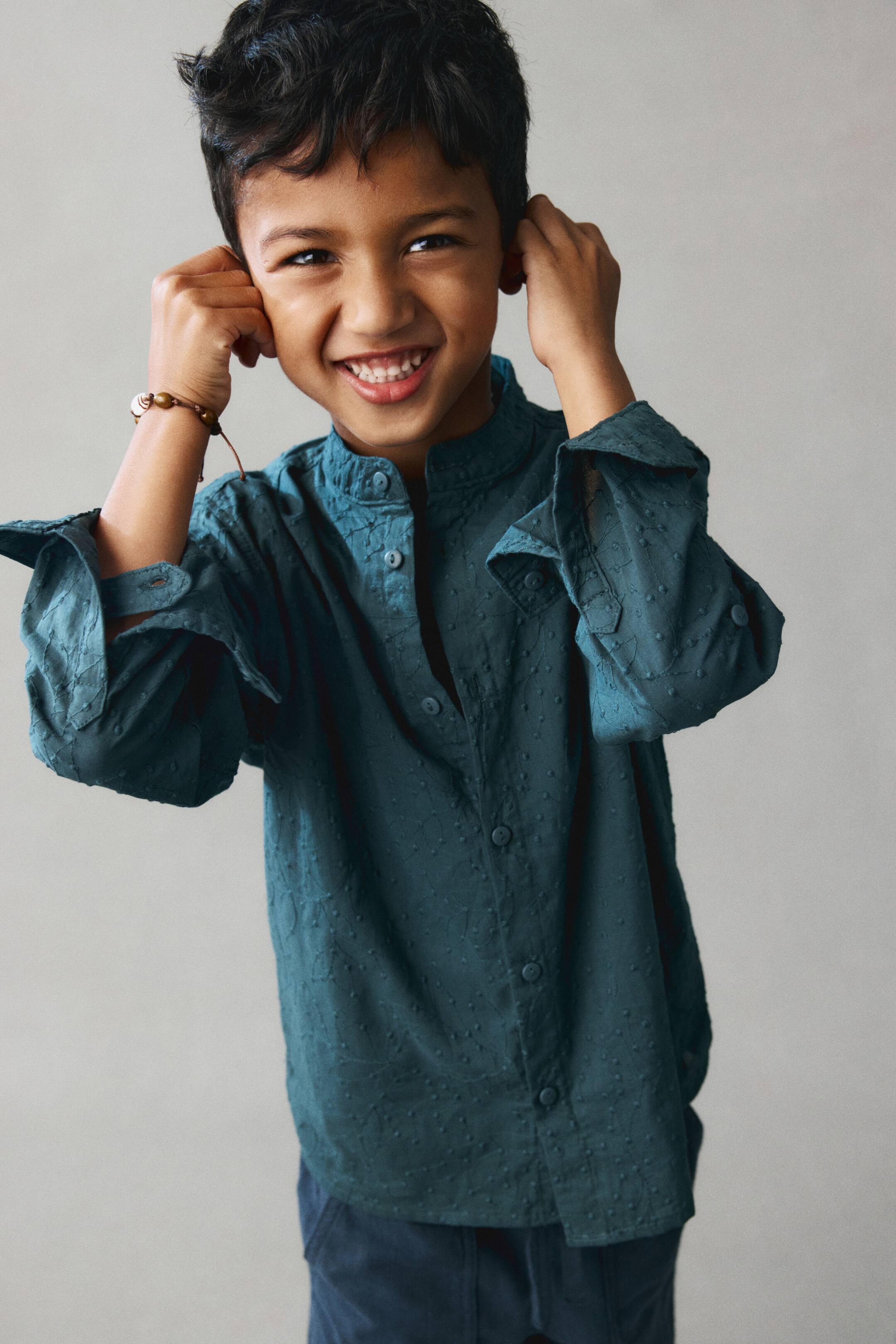 View larger image: A dark teal grandad shirt in a patterned cotton weave is shown from the front, featuring a band collar and matching buttons down the front. The long sleeves are rolled up on the arms, displaying the button cuffs and button tabs.