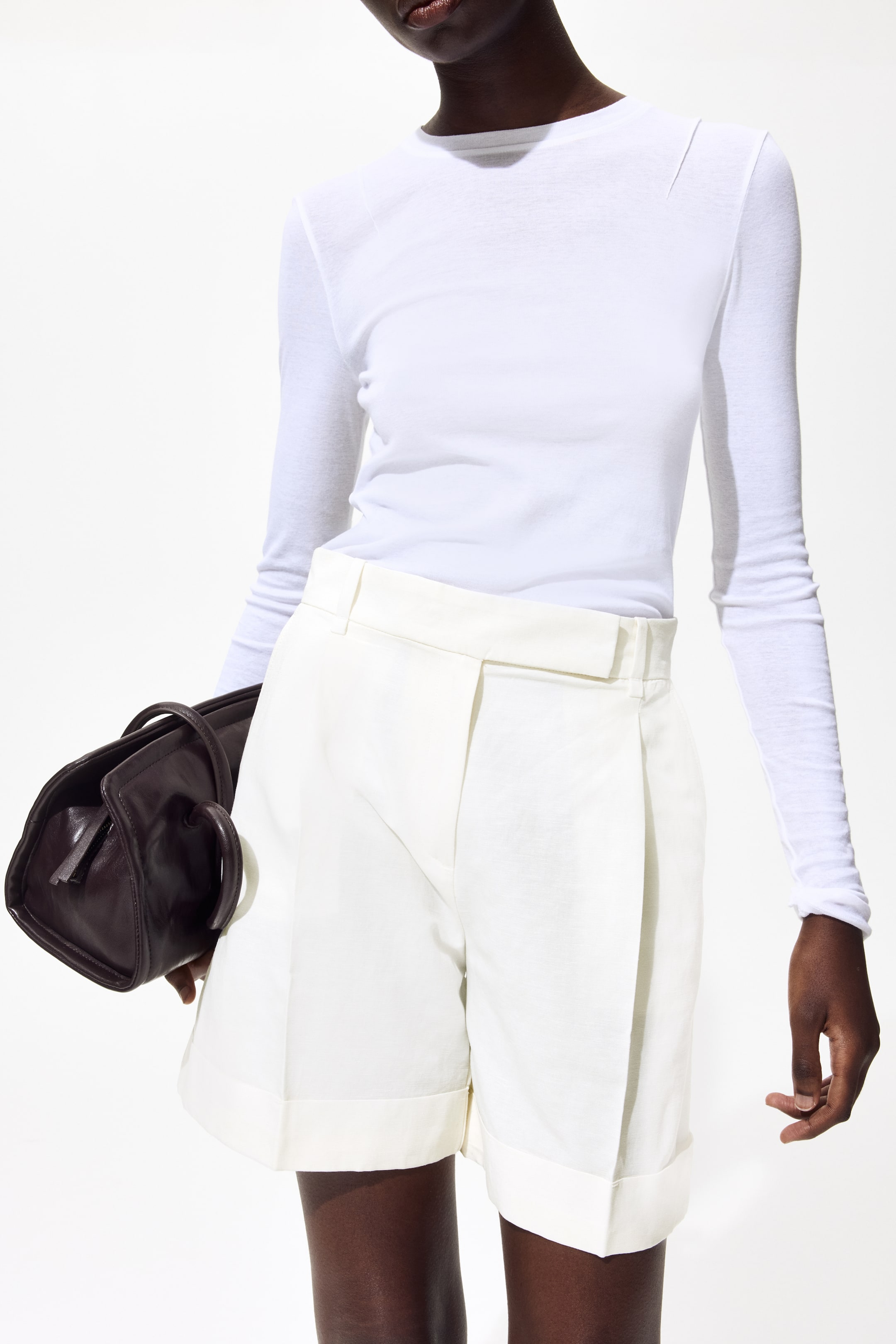 拡大画像を表示: 白い長袖トップスは、クルーネックと左肩の斜めの縫い目が特徴で、白いプリーツショーツの上に重ねられています。ショーツは折り返しの裾とベルトループを備え、両方の衣服が前を向いています。