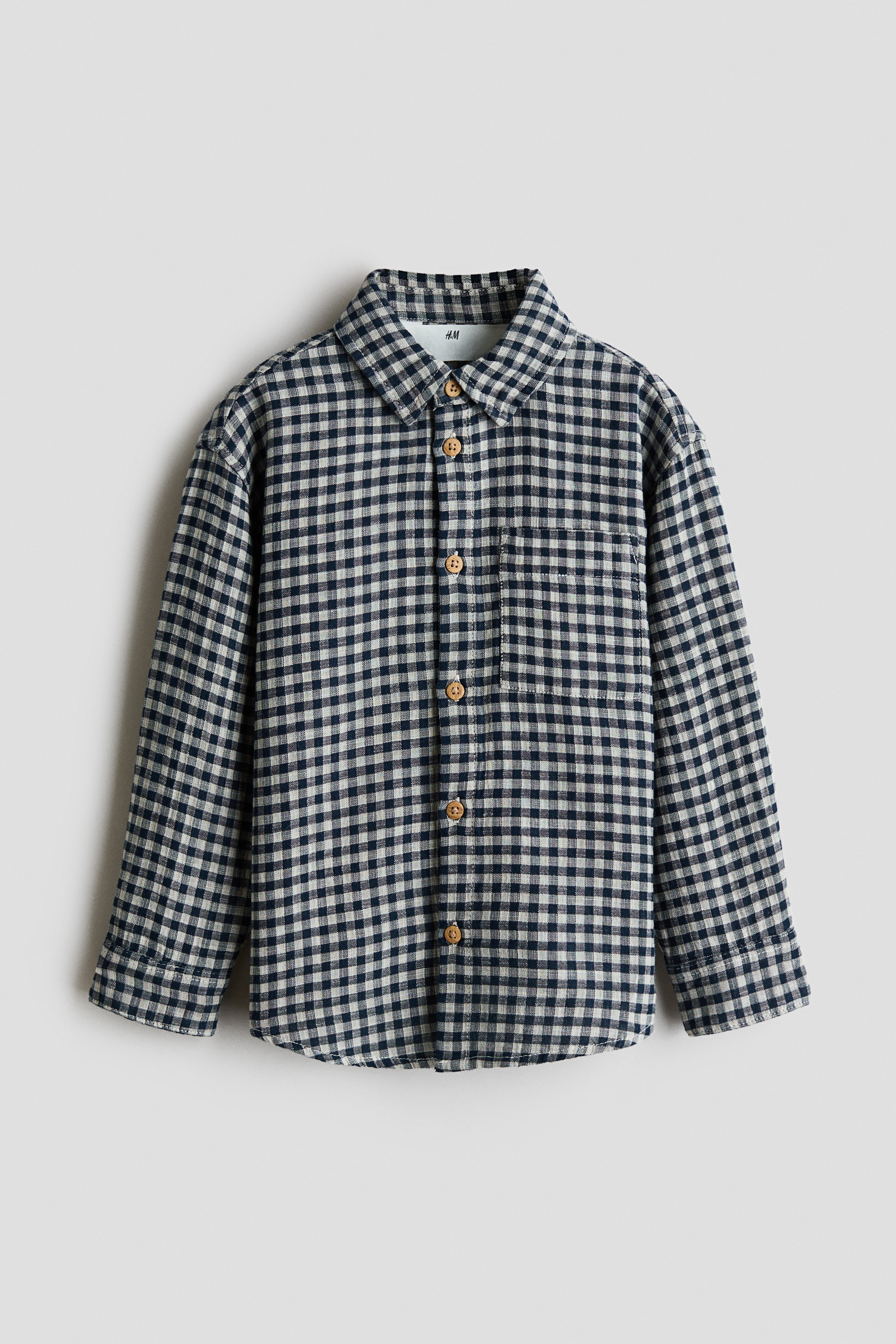 Een losvallend katoenen mousselineshirt in een marineblauw en wit gingham-ruitpatroon wordt open gedragen over een wit T-shirt, waardoor de ontspannen valling en de omgeslagen kraag mooi tot hun recht komen. Afgebeeld vanuit vooraanzicht, heeft het shirt lange mouwen met manchetten met knopen en contrasterende lichtbruine knopen langs de knoopsluiting aan de voorkant.