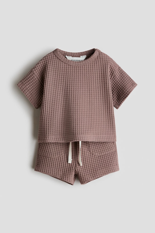 A subtle brown waffled jersey top and shorts set is showcased from the front. The top highlights a round, trimmed neckline with a press-stud on one side, dropped shoulders, and short sleeves, while the shorts come with a drawstring waist and patch front pockets. - 1