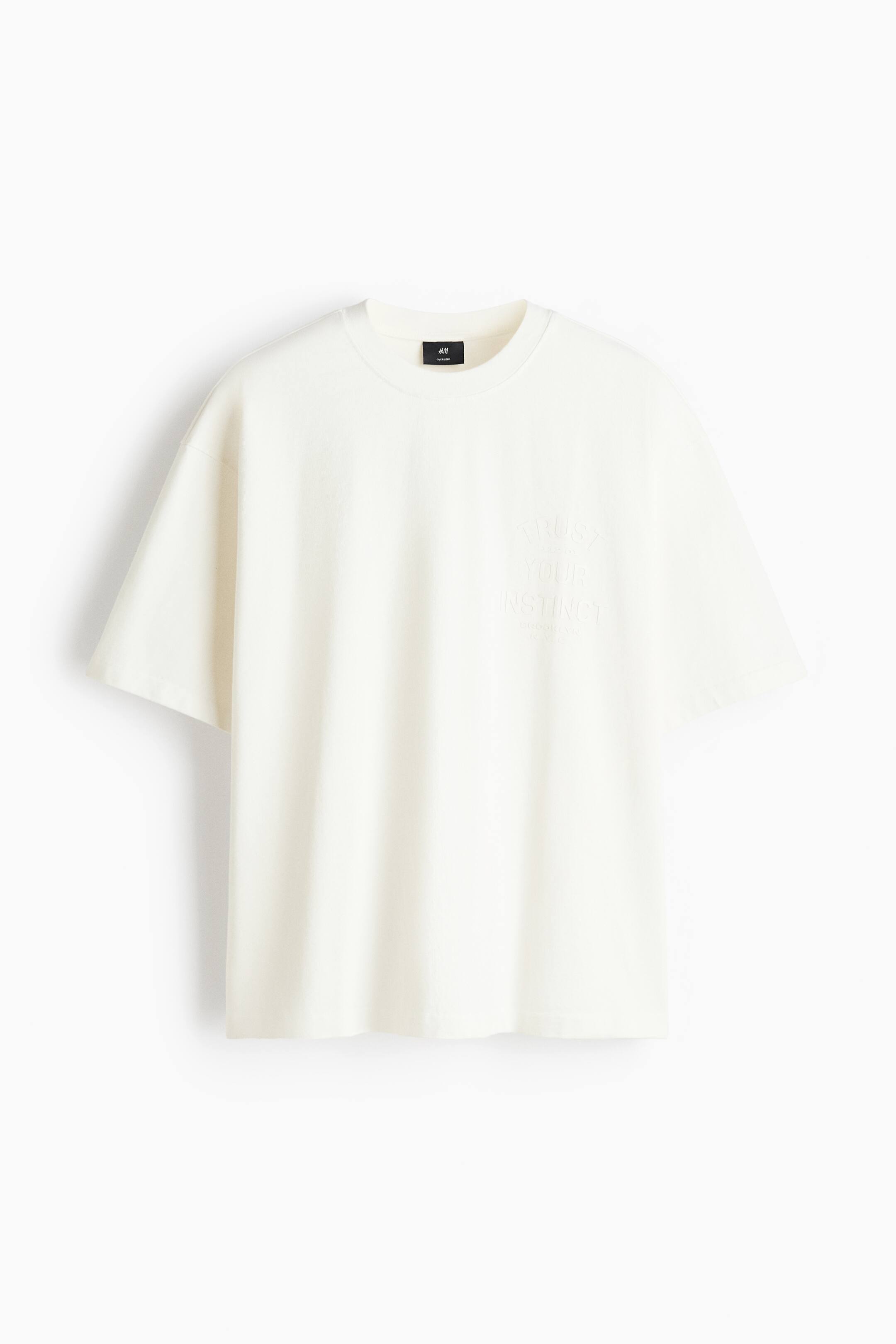 Voir une image plus grande: Un t-shirt surdimensionné blanc cassé est présenté de face, avec un col rond côtelé, des épaules tombantes et des manches courtes larges. Le t-shirt met en valeur un graphisme textuel discret en relief ton sur ton sur la poitrine gauche, indiquant « TRUST YOUR INSTINCT ».
