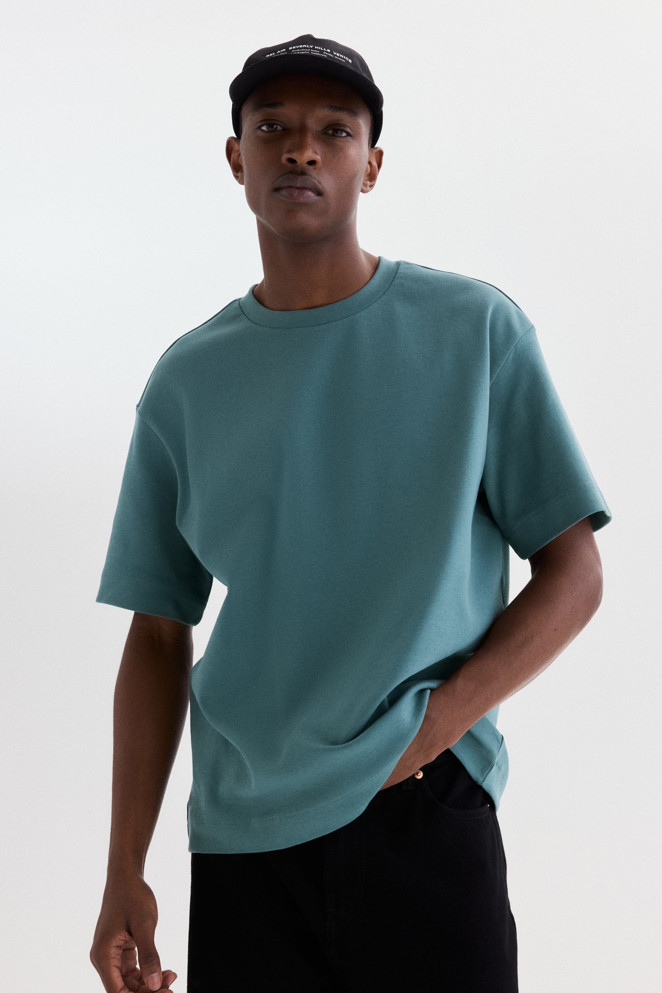 拡大画像を表示: ダークティールの半袖Tシャツが前向きに表示されており、クルーネック、ドロップショルダー、ワイドスリーブを特徴としています。リラックスフィットはストレートヘムでなめらかにドレープしており、右側で手がそのドレープに触れています。