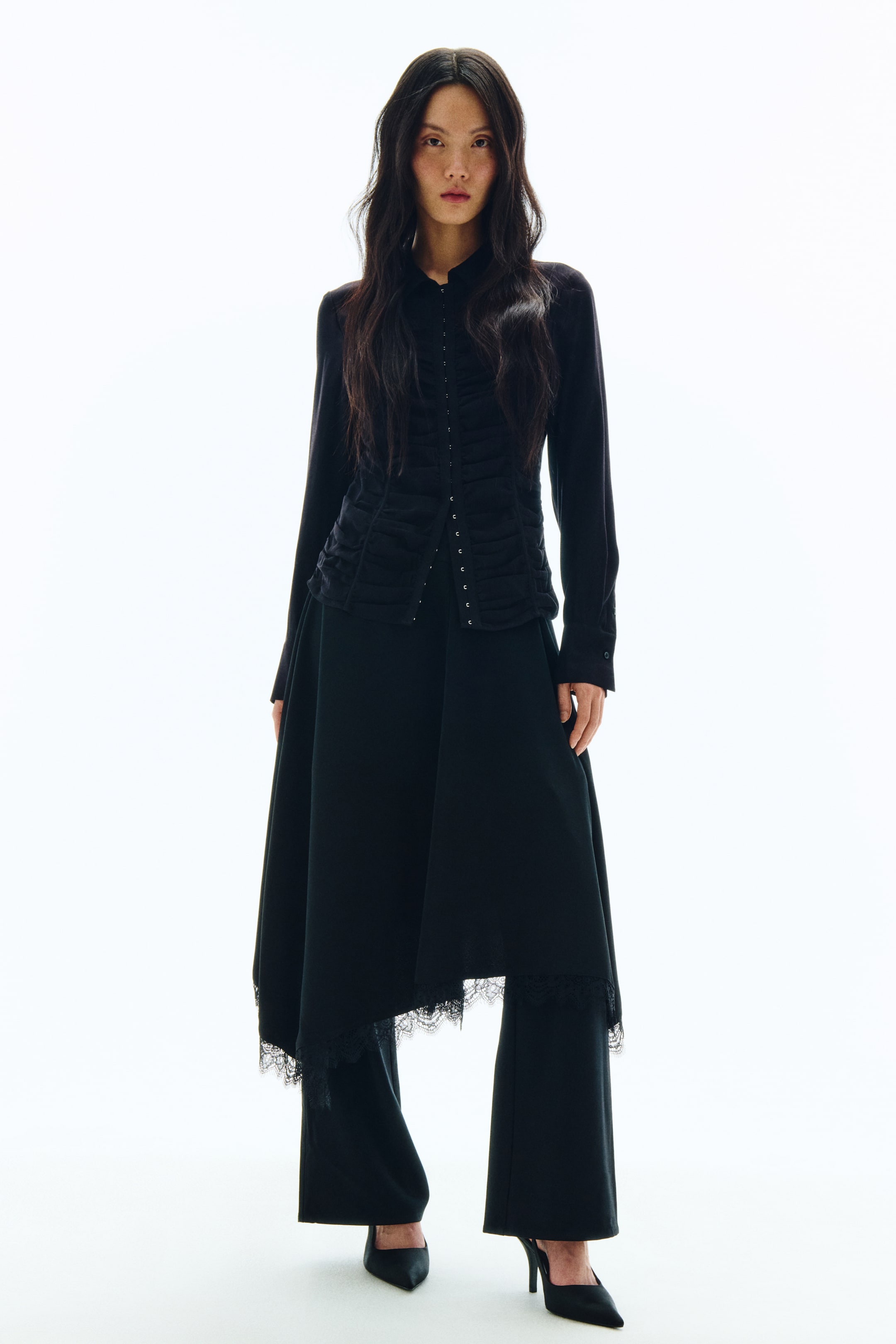 拡大画像を表示: 黒のカラー付きシャツは、フロントにギャザーを寄せたパネルとホック留めのディテールが施されています。ブラックのアシンメトリーなミディスカートは、ブラックのストレートレッグパンツの上にレイヤードされており、スカートの裾にはレーストリムがあしらわれています。