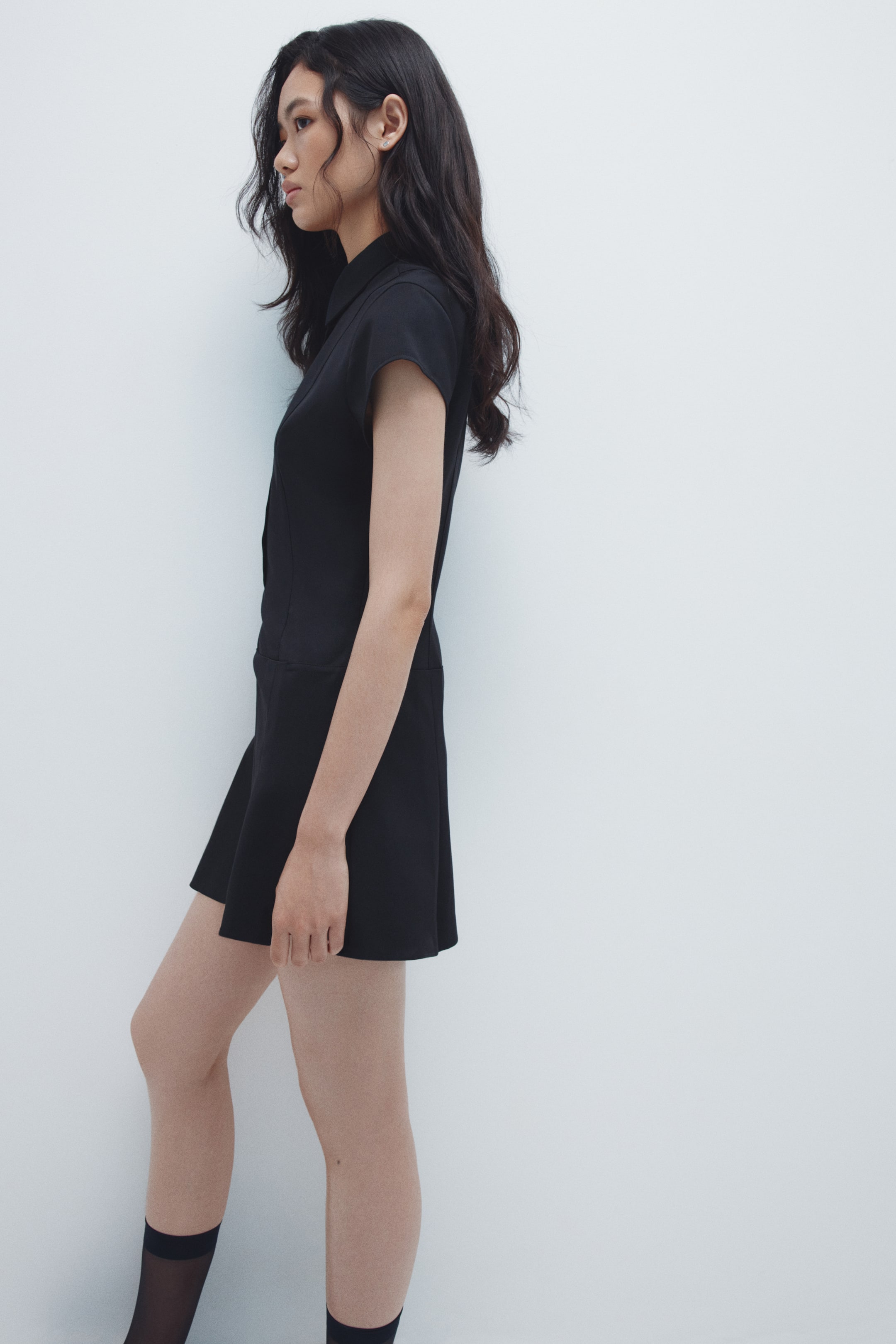 拡大画像を表示: 黒い襟付きドレスは、サイドプロファイルで紹介されています。ショートキャップスリーブ、ボタンフロントのボディス、フィットしたシルエットを強調する短く広がったスカートが特徴です。