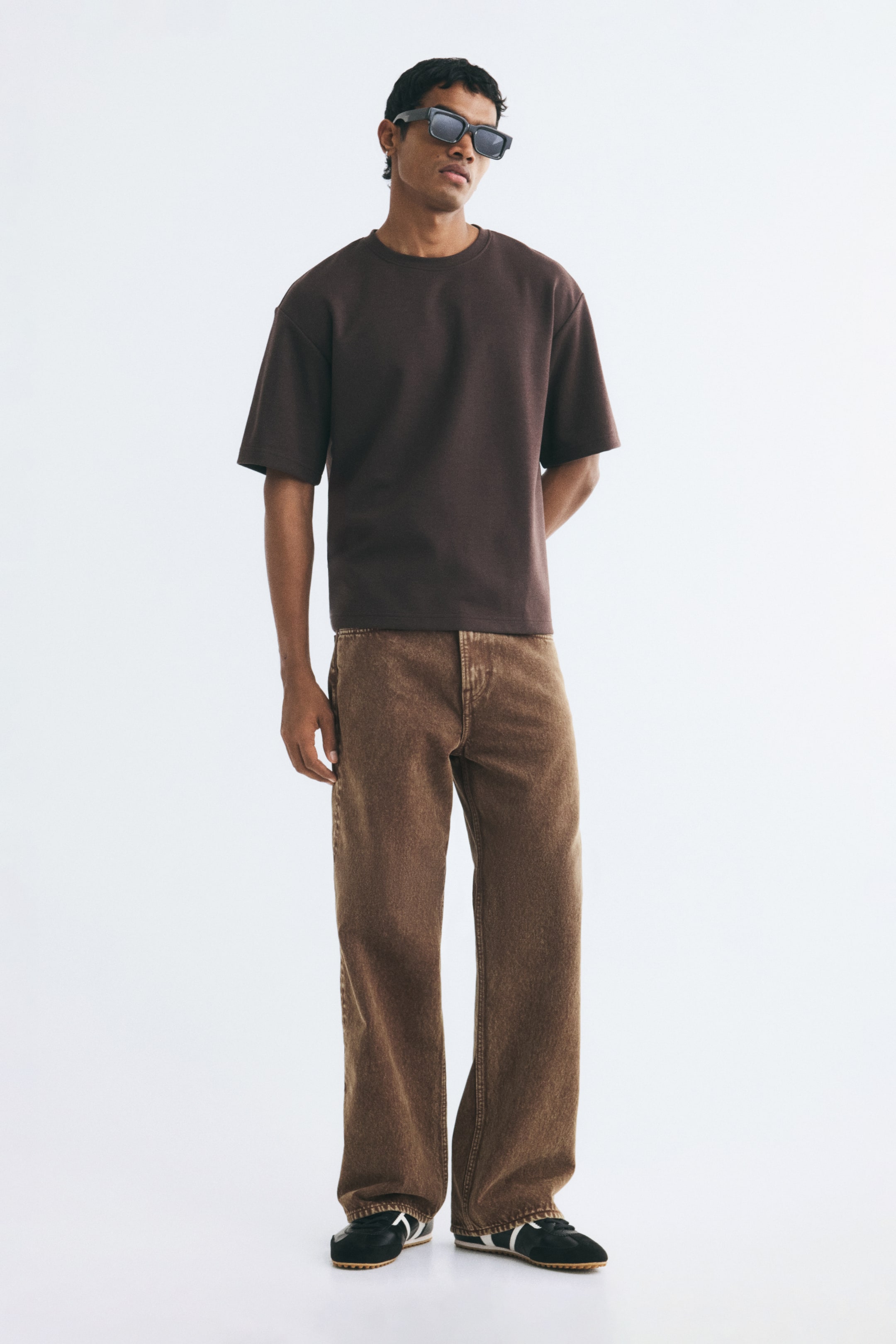 View larger image: Brown denim jeans featuring a loose fit and straight legs are worn by the model. The jeans are styled with a short-sleeved dark brown T-shirt and black sneakers.