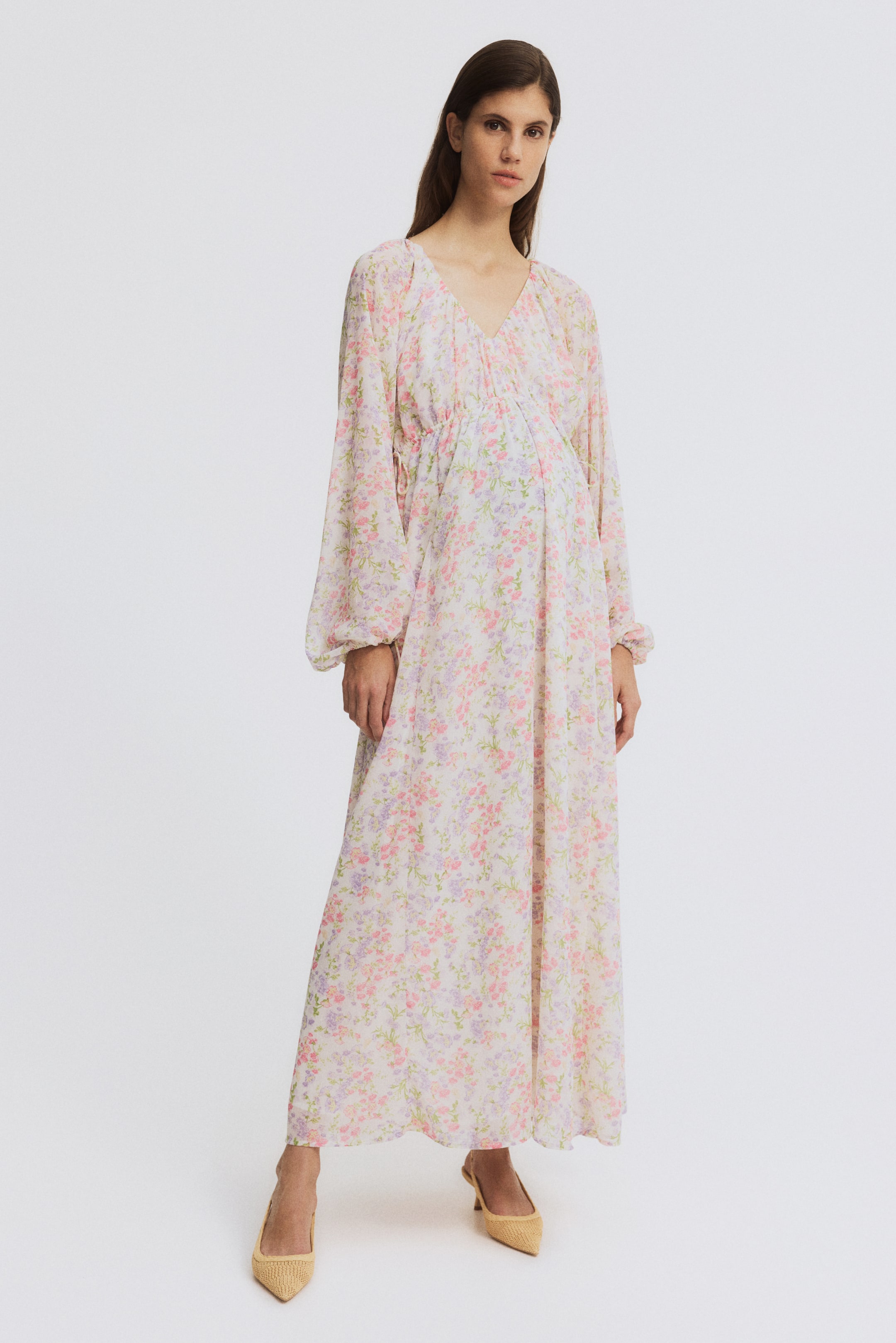 Ampliar la imagen: La modelo lleva un vestido largo y suelto de gasa arrugada, estampado con flores en tonos rosa claro y morado. Presenta un profundo escote en V, mangas largas raglán abullonadas con puños elásticos, finos cordones que se atan debajo del busto a los lados, y está forrado en la parte superior.