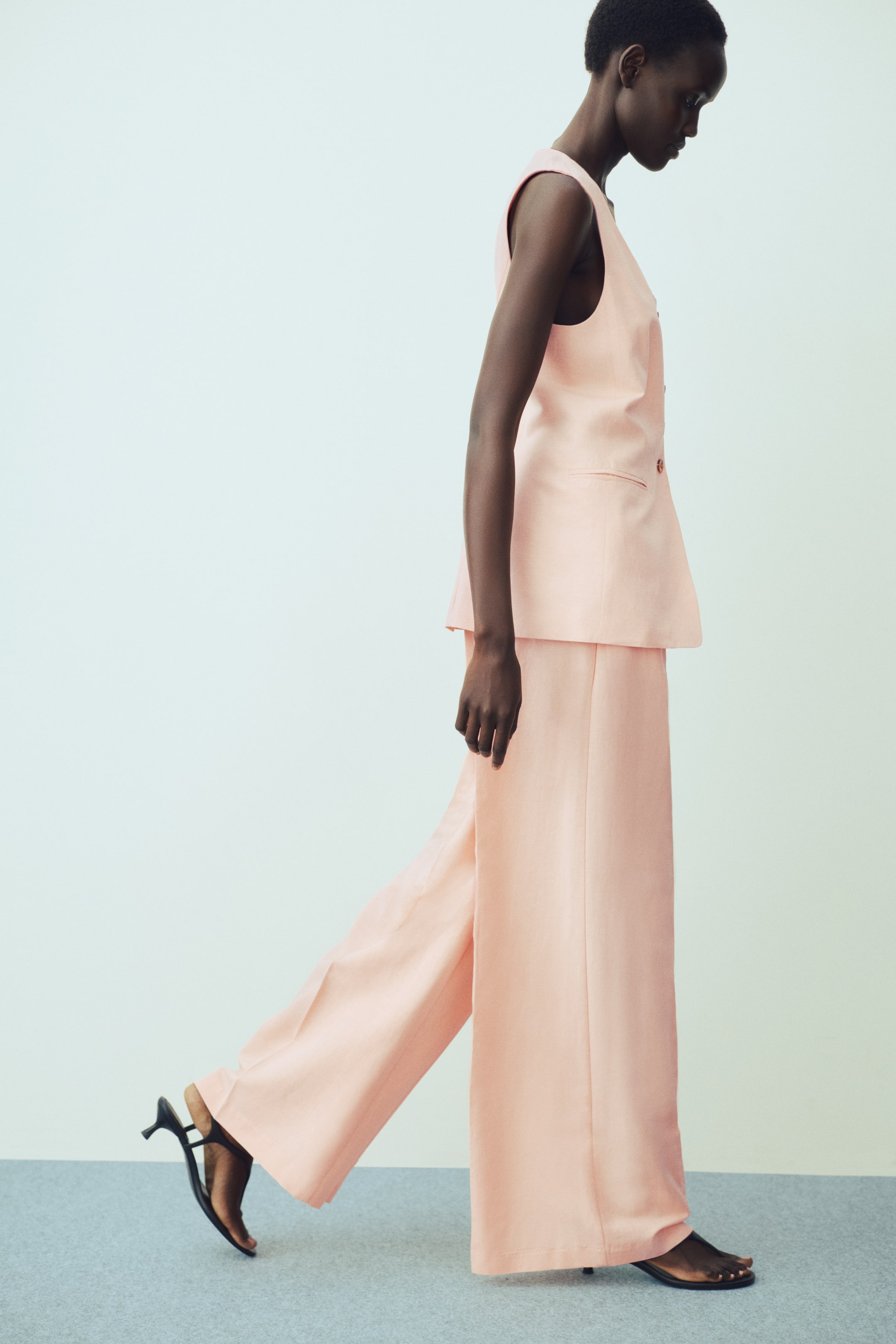 View larger image: The light peach sleeveless vest and matching wide-leg trousers are showcased from a side profile. The vest features a V-neckline, two visible buttons, and a pocket, while the trousers flow broadly, partially covering black high-heeled sandals.