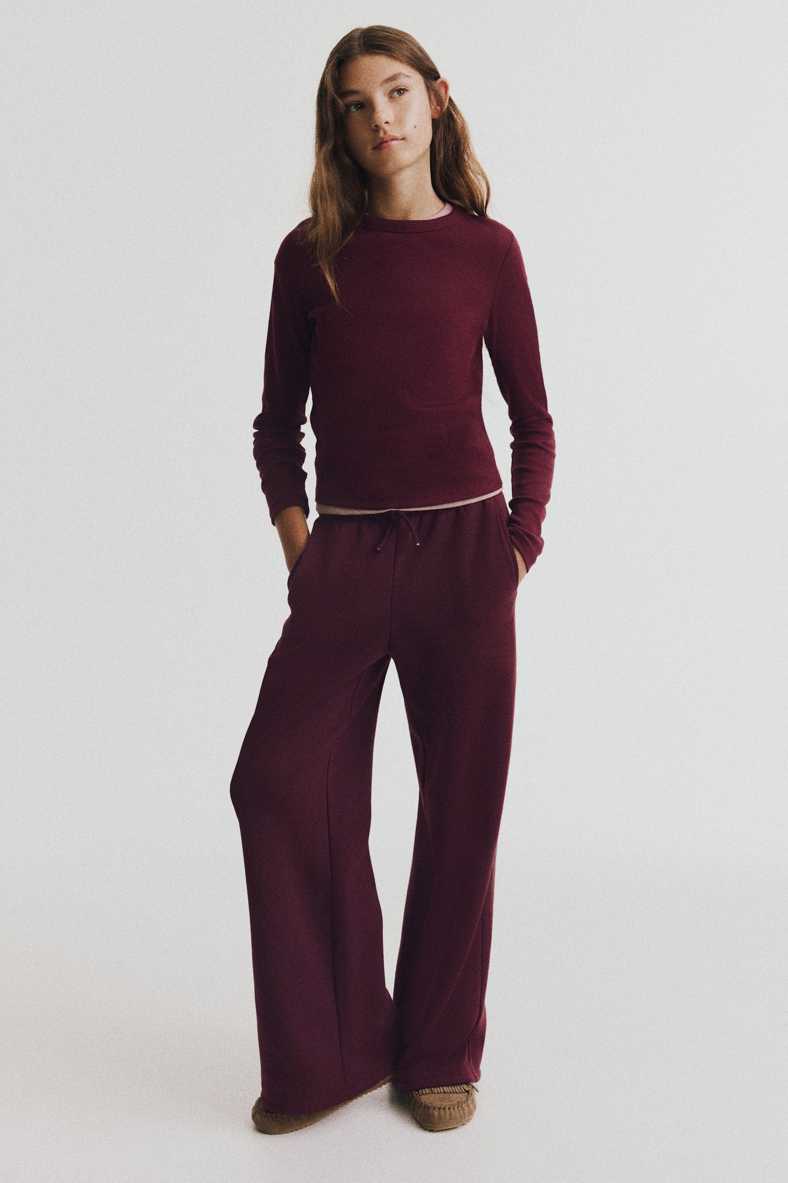 A long-sleeve, fitted top in burgundy ribbed cotton jersey is shown from the front, featuring a round neckline and a straight-cut hem. A contrasting light pink underlayer is peeking out at the collar and bottom hem. - 1