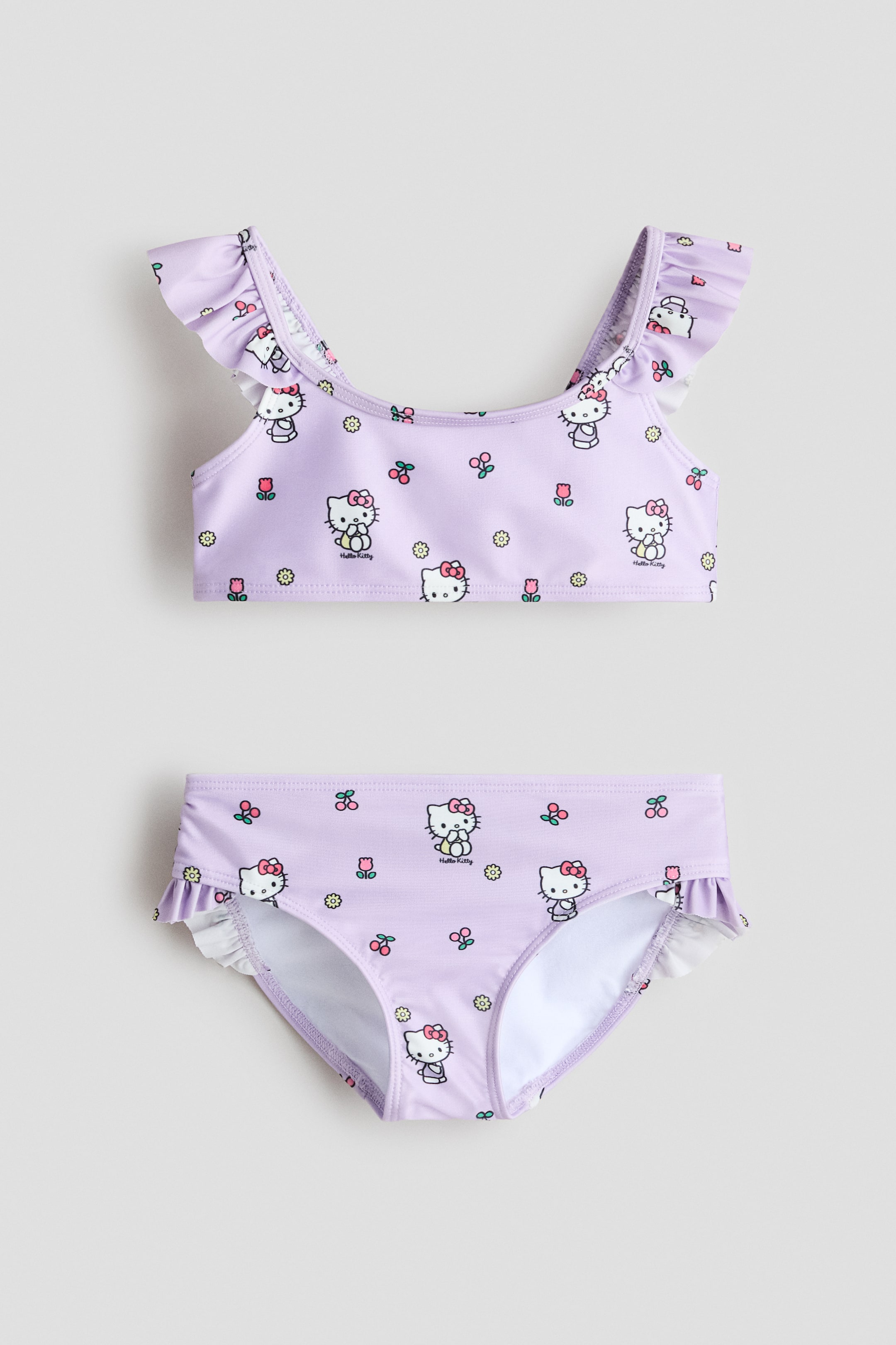 Προβολή μεγαλύτερης εικόνας: Σετ μαγιό δύο τεμαχίων για παιδιά σε απαλό μωβ χρώμα, με επαναλαμβανόμενο μοτίβο λευκών χαρακτήρων Hello Kitty με ροζ φιόγκους, διακοσμημένο με μικρά λουλούδια και κεράσια. Το τοπ του μπικίνι διαθέτει τιράντες ώμων με βολάν, ενώ το αντίστοιχο σλιπ έχει βολάν περιμετρικά στις ανοίξεις των ποδιών — όλα φαίνονται από μπροστά.