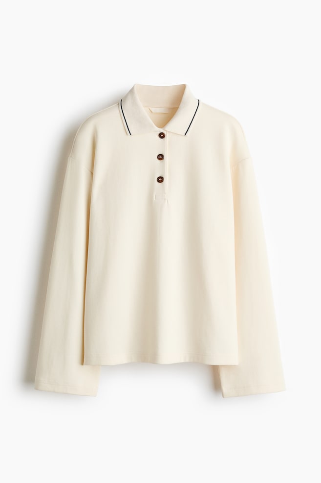 A cream long-sleeved polo shirt is displaying a pointed collar edged with a thin black stripe, a three-button placket with dark brown buttons, and a relaxed fit with long sleeves and a straight hem. - 1