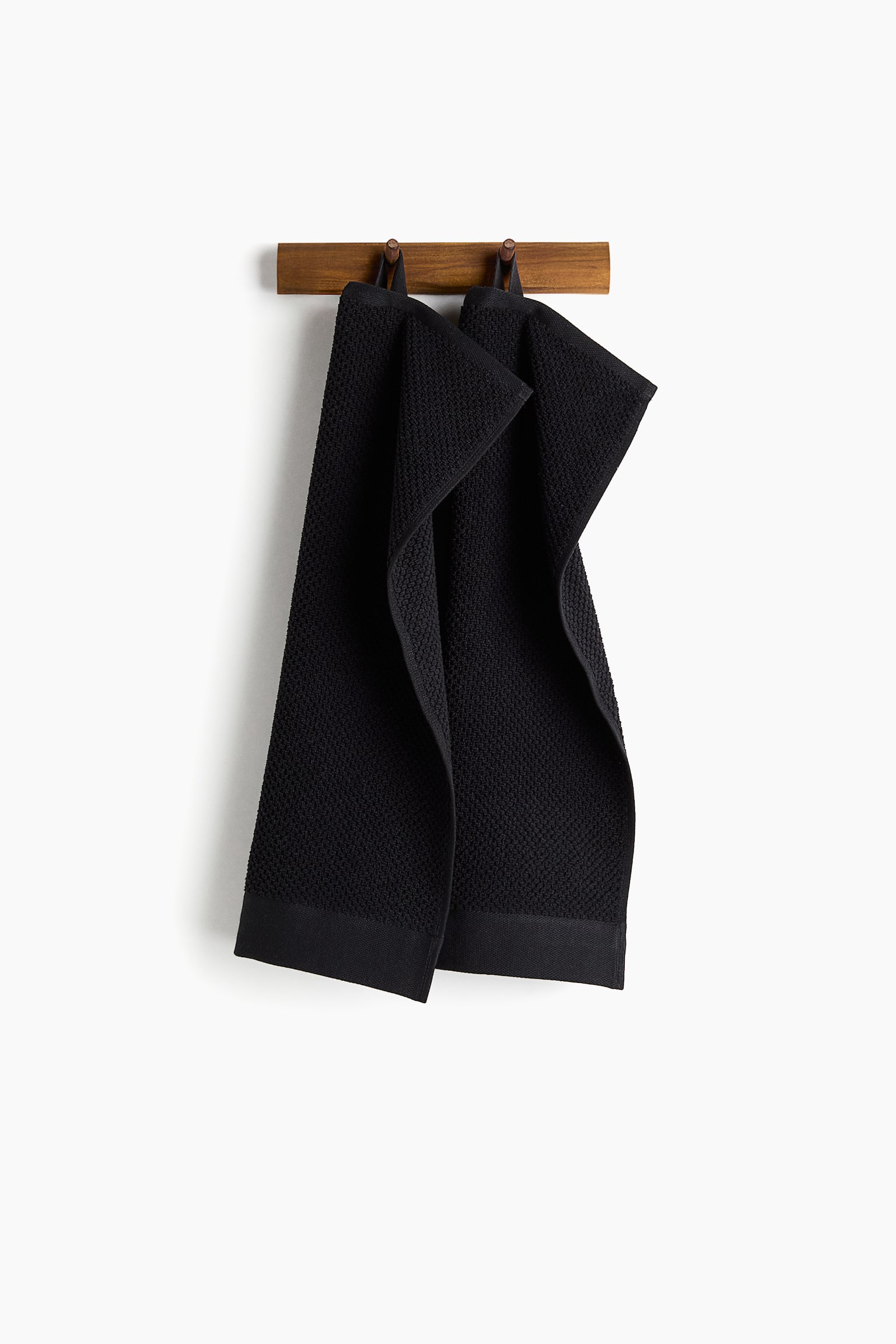 Two black towels hang side-by-side from a wooden rack. Each towel features a textured waffle weave throughout its body, a plain hemmed band at the bottom, and a fabric loop at the top for hanging. - 1