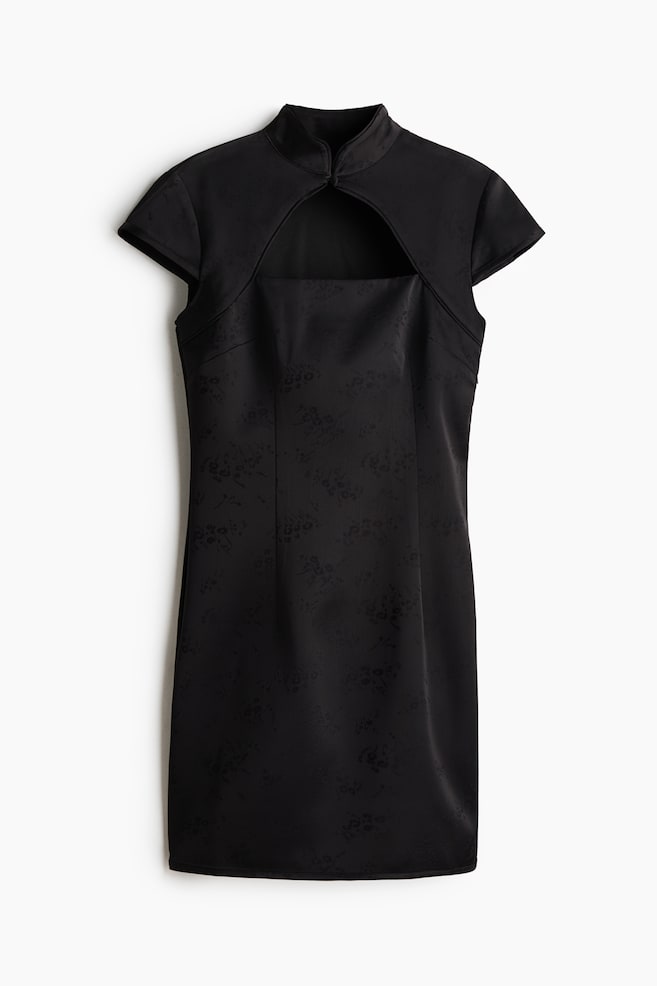 Un vestido corto y ajustado de color negro, en tejido jacquard, se muestra de frente y luce un cuello mao con cierre frontal, una gran abertura en el pecho y mangas tipo capa. La prenda presenta un sutil motivo floral con pinzas verticales que definen la cintura. - 1