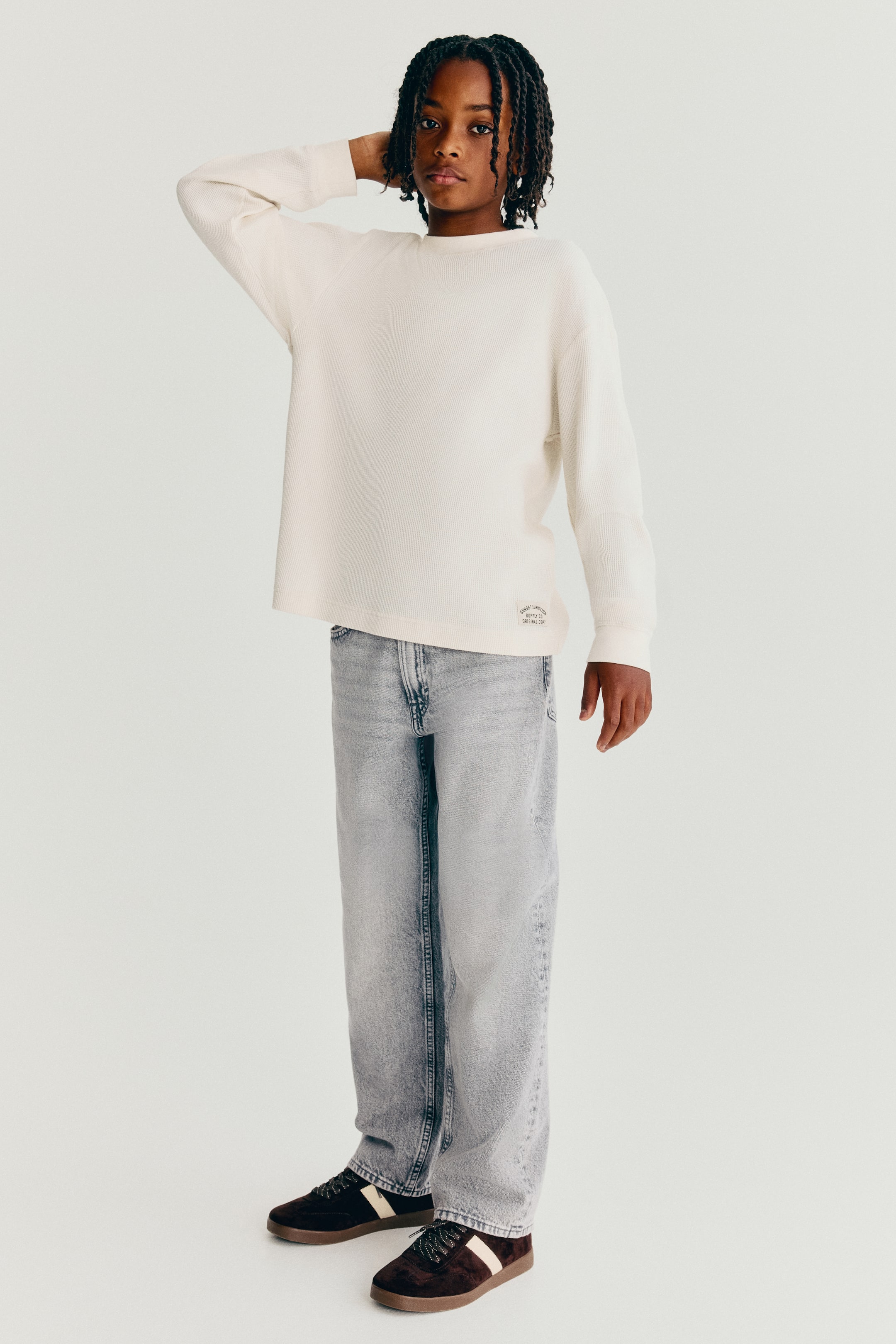 View larger image: Loose-fit, light-wash denim jeans are shown from the front, featuring a relaxed silhouette with straight legs that drape over dark trainers. The classic five-pocket jeans are styled with an off-white long-sleeve top that partially covers the waistband.