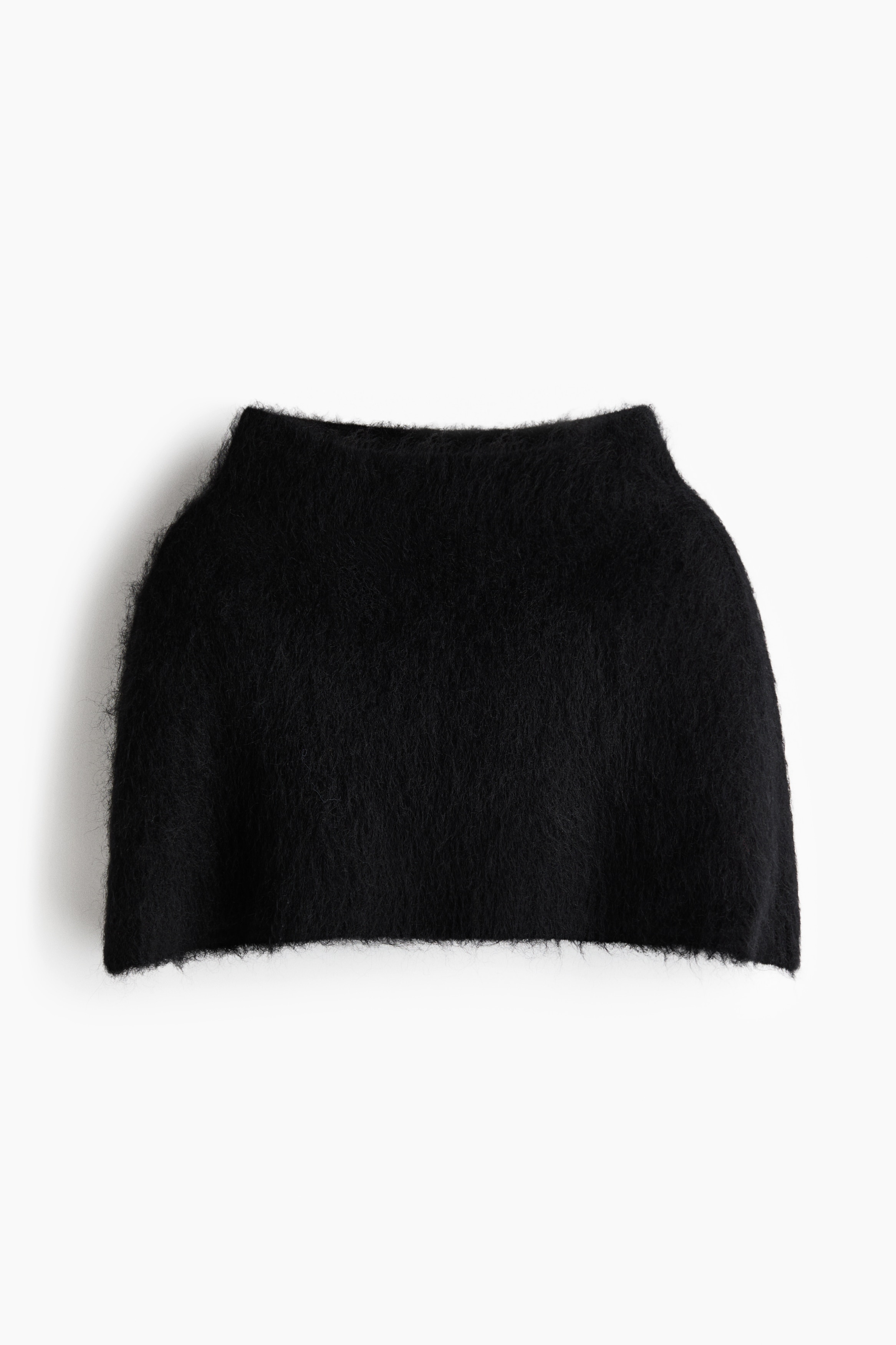 Une cape noire en maille cÃ´telÃ©e, en mÃ©lange de mohair et dotÃ©e dâun col montant, est portÃ©e par le mannequin. La cape texturÃ©e couvre les Ã©paules et est superposÃ©e Ã  un blazer noir structurÃ© et Ã  un pantalon blanc.