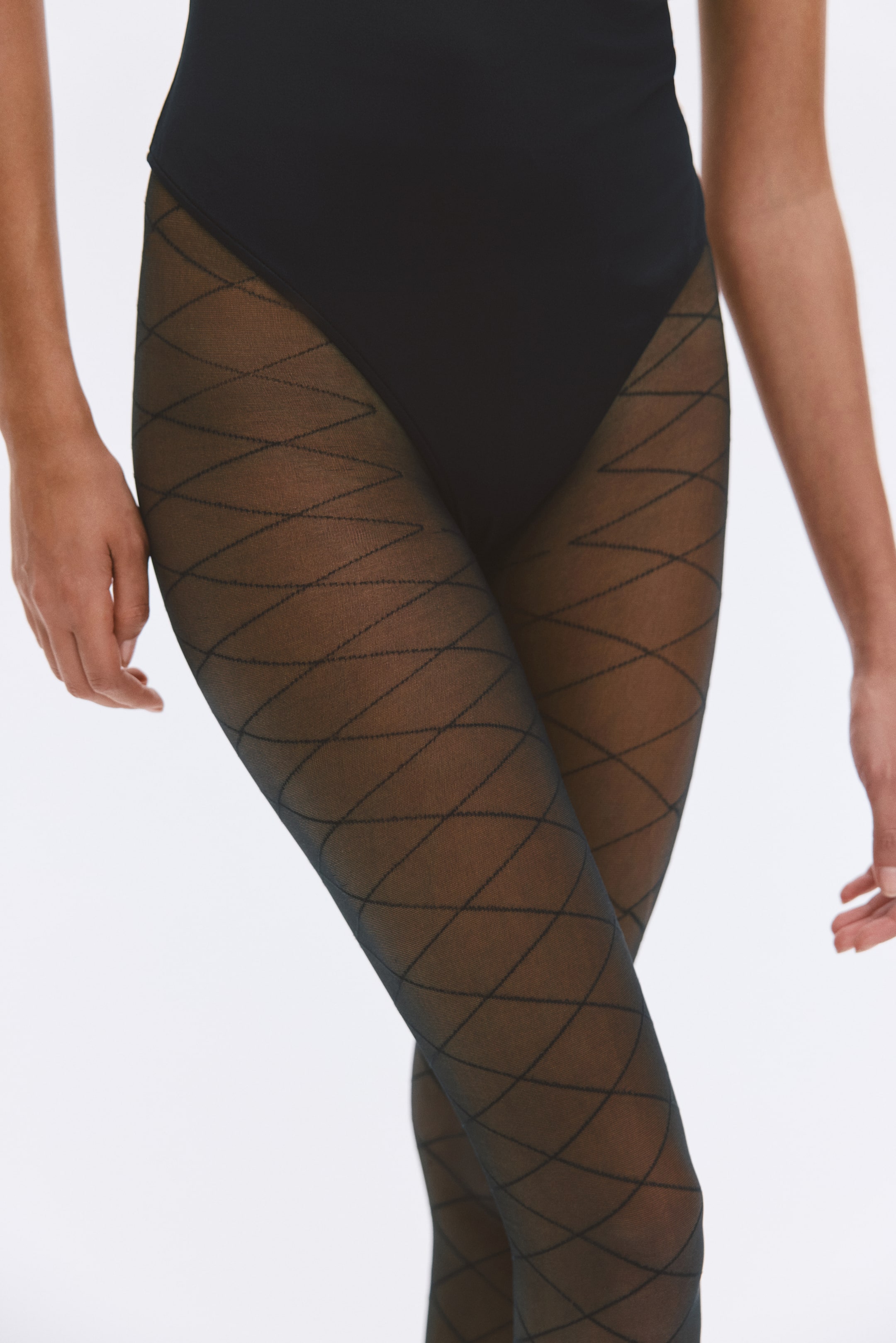 Se større billede: Gennemsigtige sorte strømpebukser med et geometrisk kryds-og-tværs-mønster bæres af modellen. Strømpebukserne er stylet under en sort bodystocking med høj benudskæring.