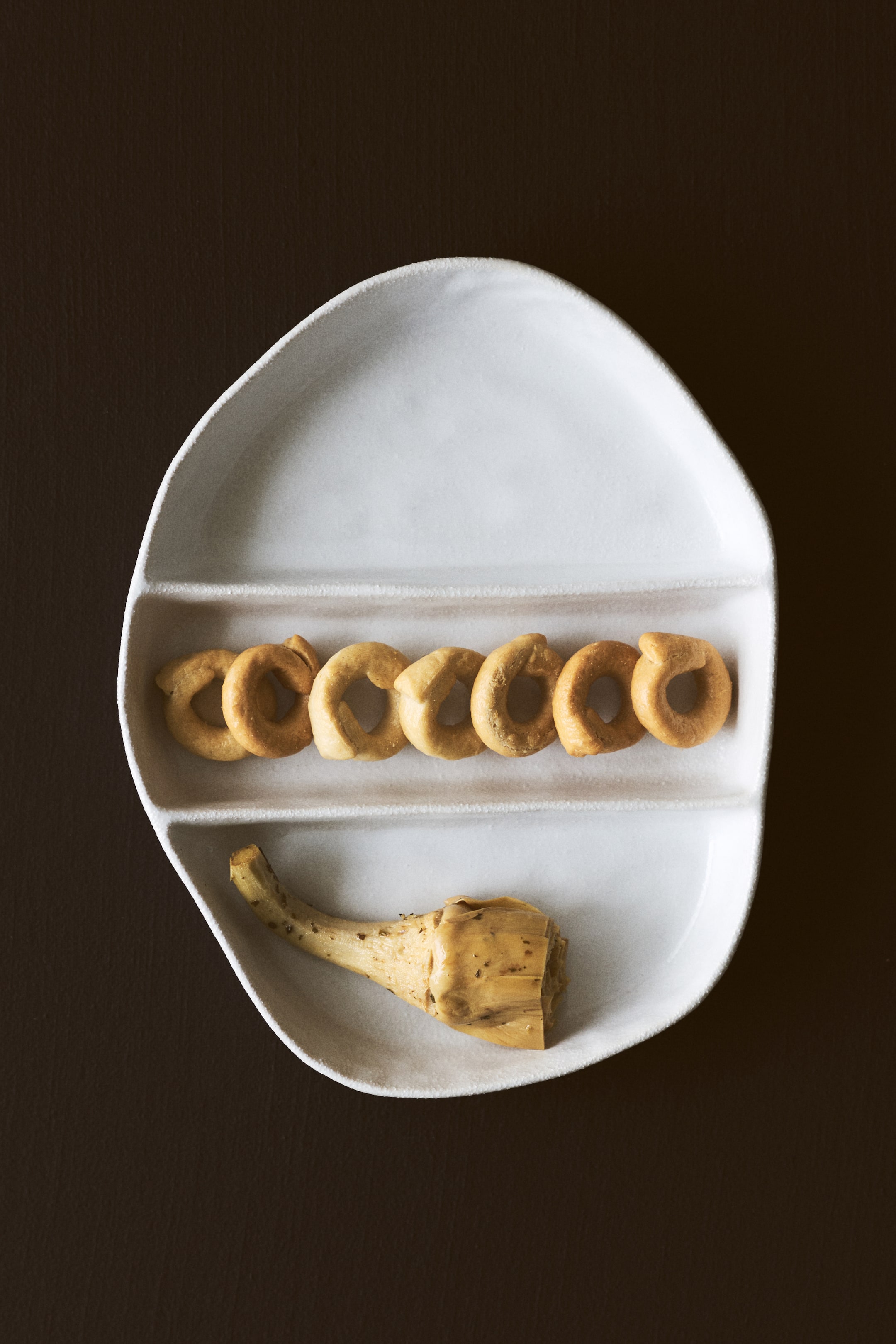 이미지 확대 보기: 흰색의 불규칙한 모양을 가진 서빙 접시가 위에서 내려다본 각도로 보입니다. 세 개의 길고 좁은 칸이 특징입니다. 가운데 칸에는 링 모양의 비스킷이 줄지어 있으며, 아래 칸에는 아티초크 하트가 하나 놓여 있습니다.