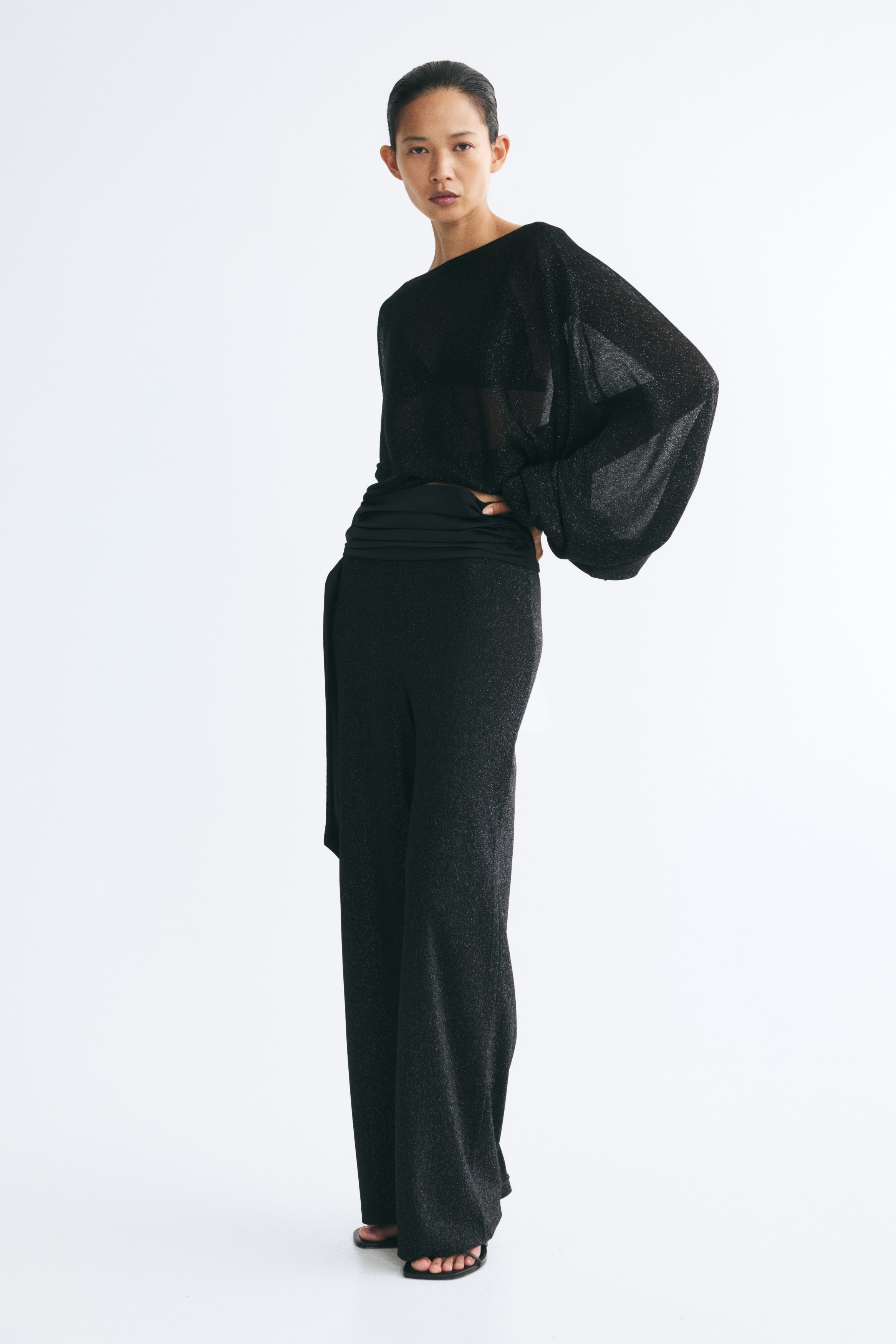 A black oversized knitted top featuring glittery threads is hanging in a front view, showcasing a boat neckline and draped, voluminous cape sleeves tapering into narrow elasticated cuffs.