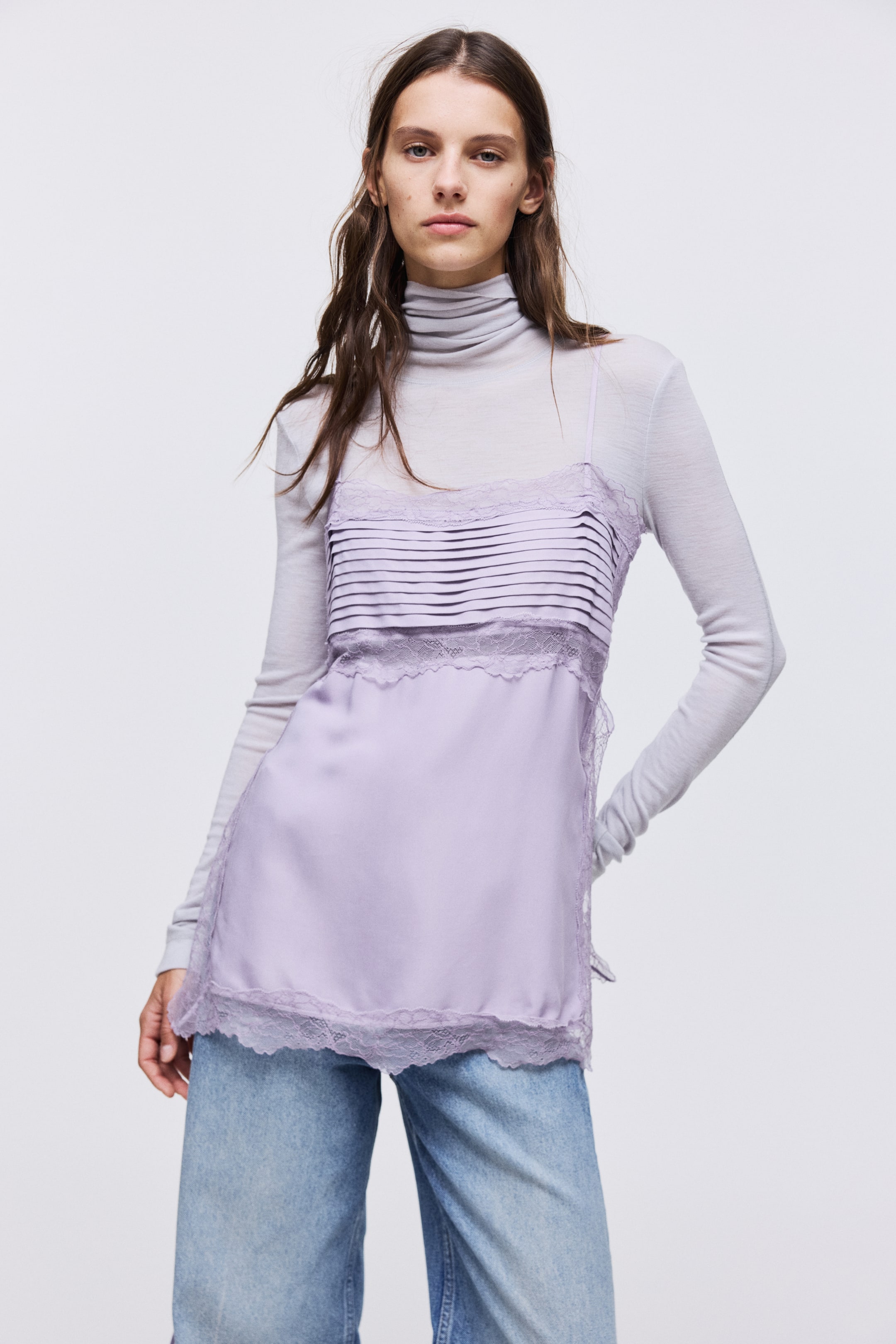 View larger image: A lavender long top, made of a viscose weave with lace edges and insets, is being worn by the model. It is featuring a pleated section over the bust, adjustable spaghetti shoulder straps, and a high slit with narrow ties at each side, layered over a light grey turtleneck.