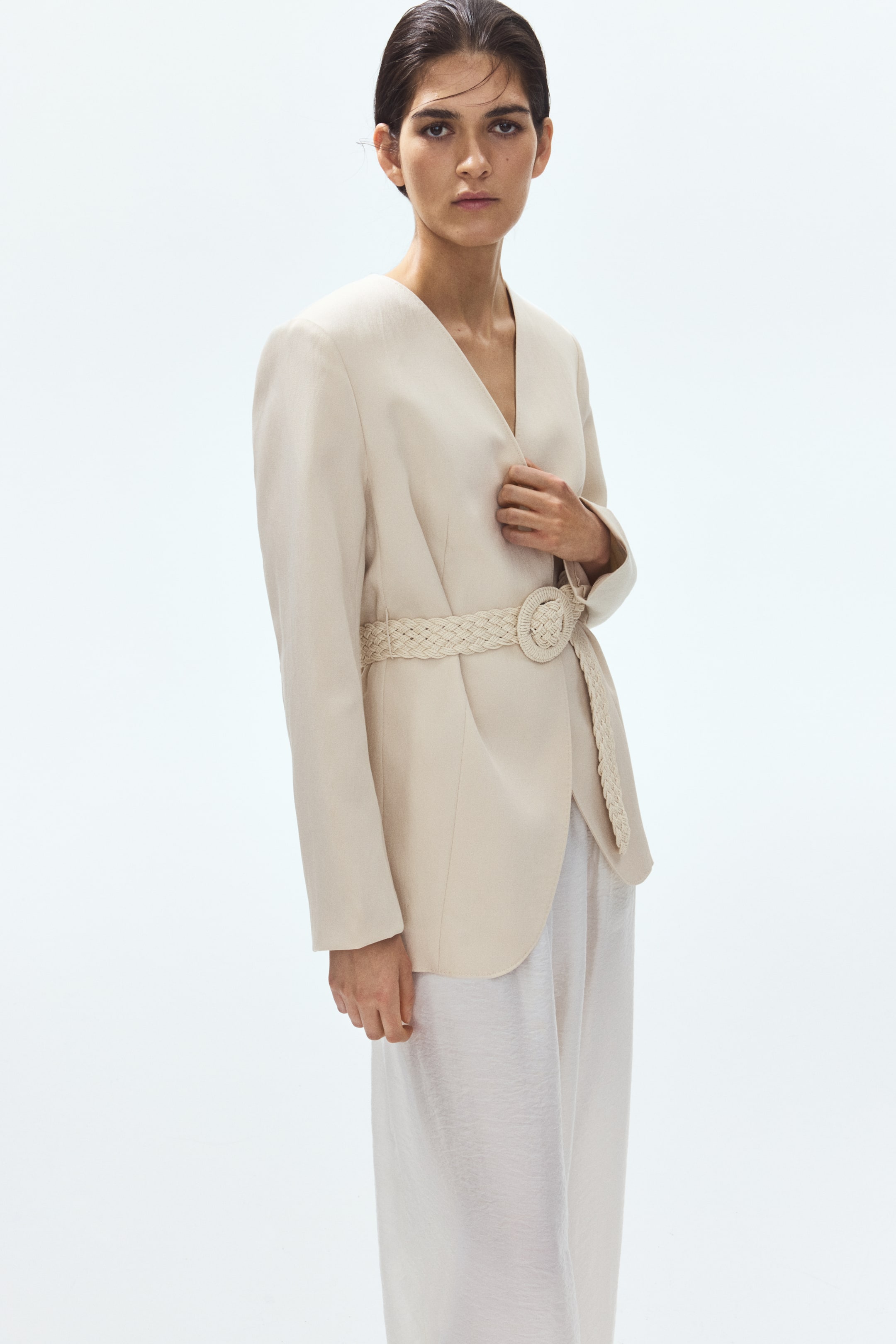 Visualizza immagine più grande: Un blazer beige chiaro senza collo è rivolto in avanti, leggermente inclinato a sinistra, mettendo in risalto lo scollo a V e le spalle strutturate mentre arriva fino alla parte superiore della coscia. È stretto in vita da una cintura intrecciata abbinata con una grande fibbia rotonda, ed è indossato sopra pantaloni chiari e testurizzati.