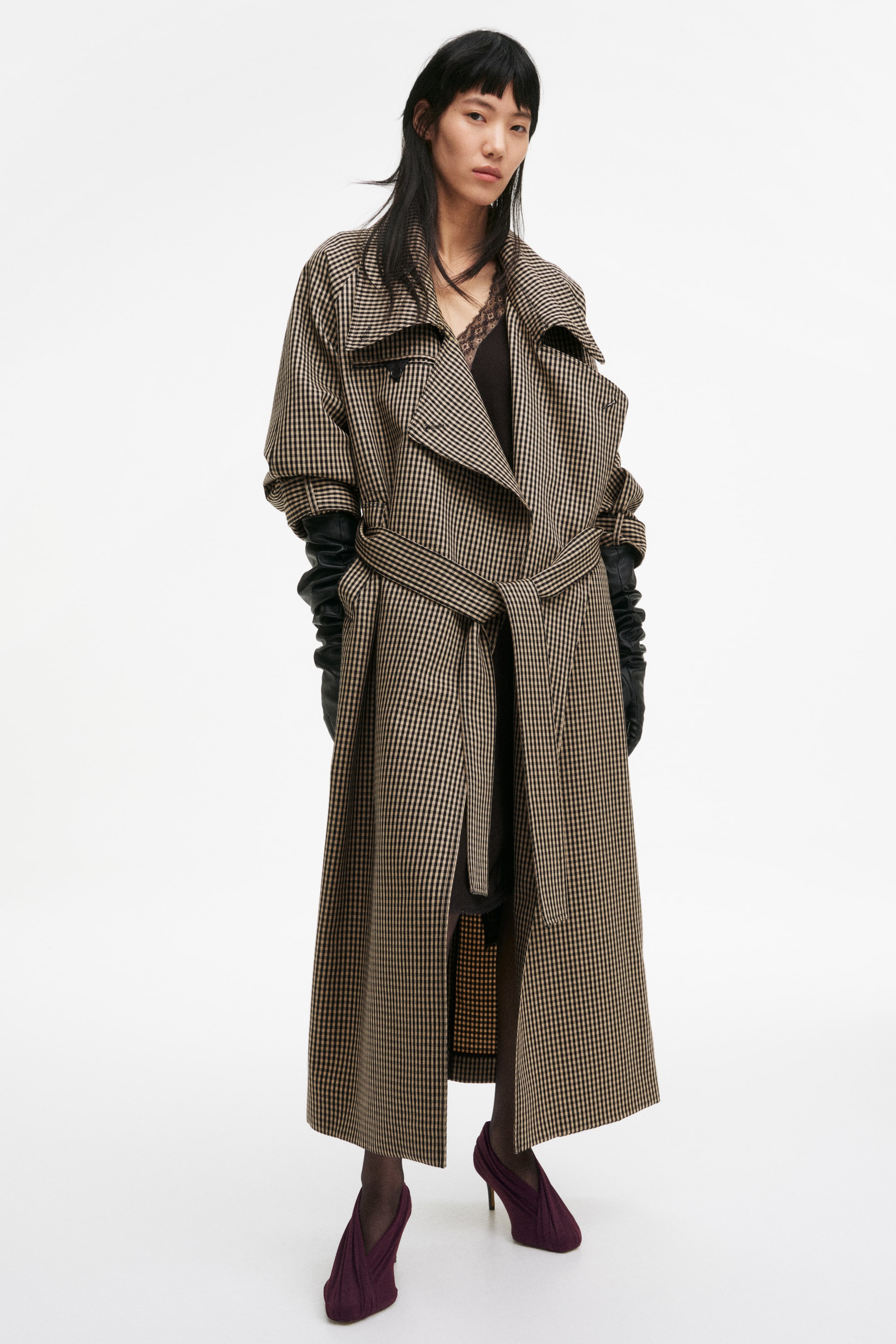 View larger image: A long trench coat in a brown and tan small check pattern features wide lapels and an oversized fit, complete with a matching fabric belt tied at the waist. Its long, voluminous sleeves drape elegantly over black leather gloves.