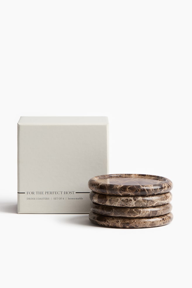 Four round brown marble drink coasters are arranged in a stack, situated next to a light beige rectangular packaging box. The coasters exhibit unique veining patterns, and the box features text on its front that reads "FOR THE PERFECT HOST DRINK COASTERS SET OF 4 brown marble". - 1