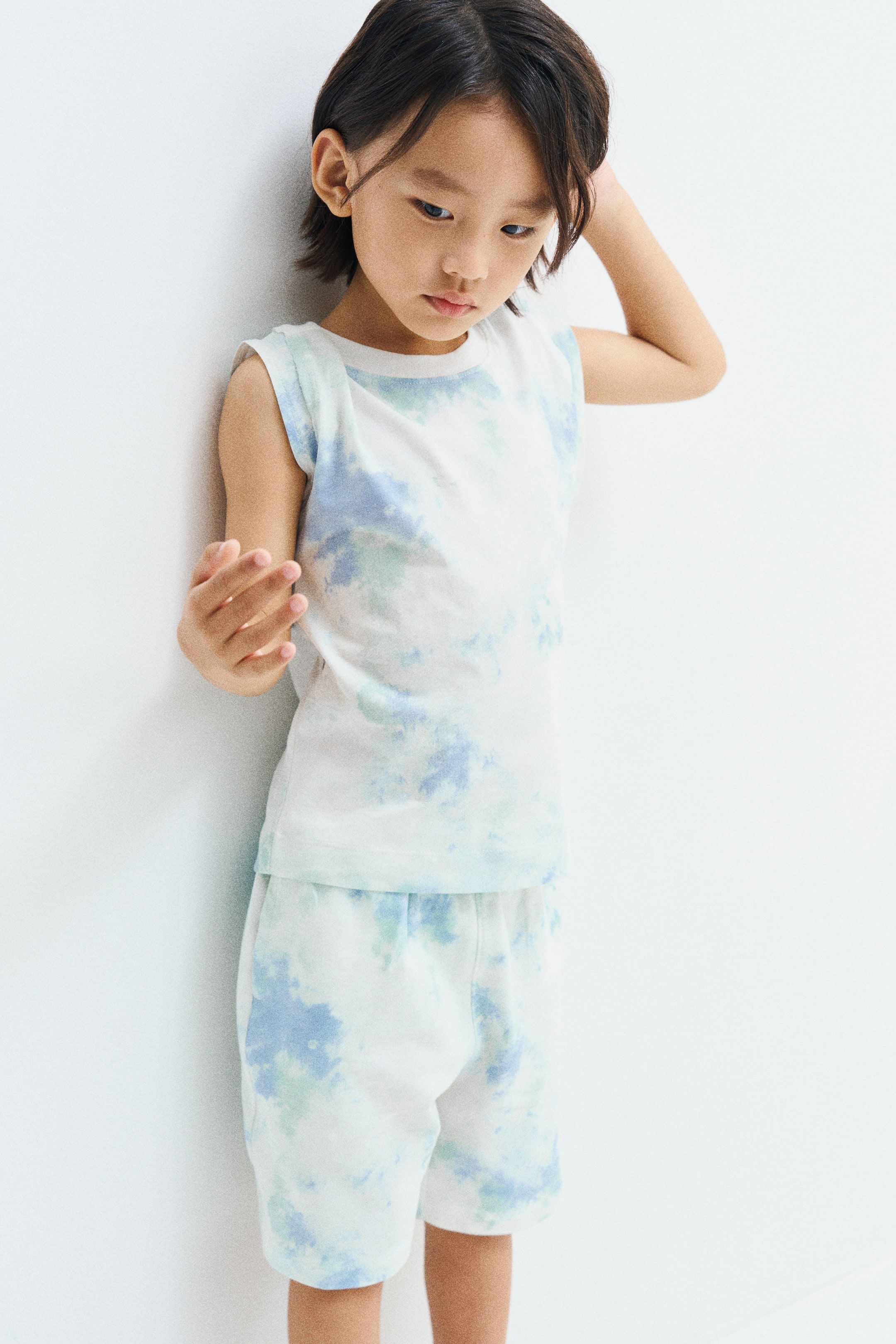 View larger image: The model is wearing knee-length shorts featuring a light blue and green tie-dye print on white fabric. The shorts are paired with a matching sleeveless top in the same print design.