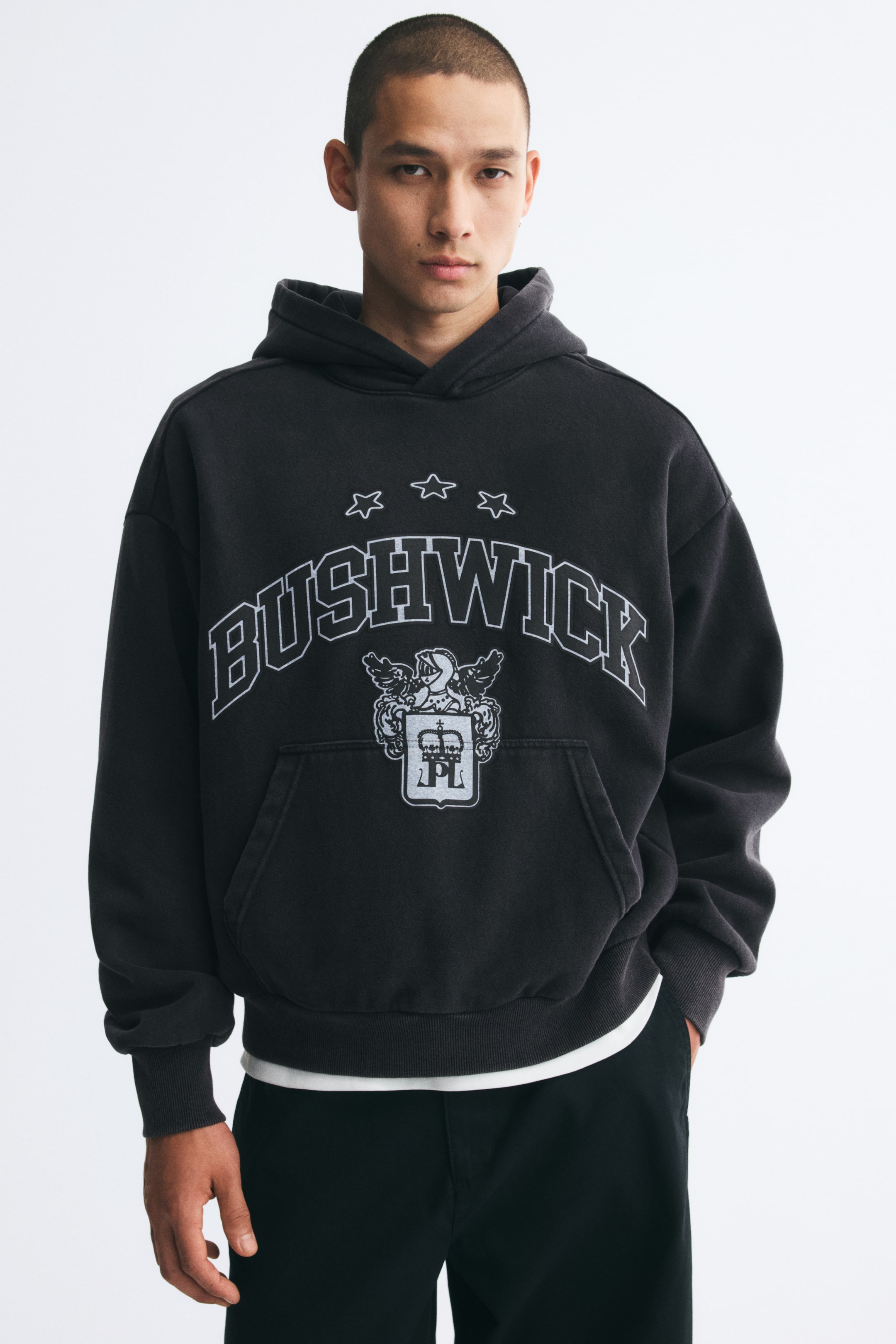 View larger image: A dark grey, oversized hoodie featuring a printed "BUSHWICK" graphic and crest on the chest is worn by the model. The hoodie has dropped shoulders, a wrap hood, a kangaroo pocket and ribbed trims, and is layered over a white T-shirt and black trousers.