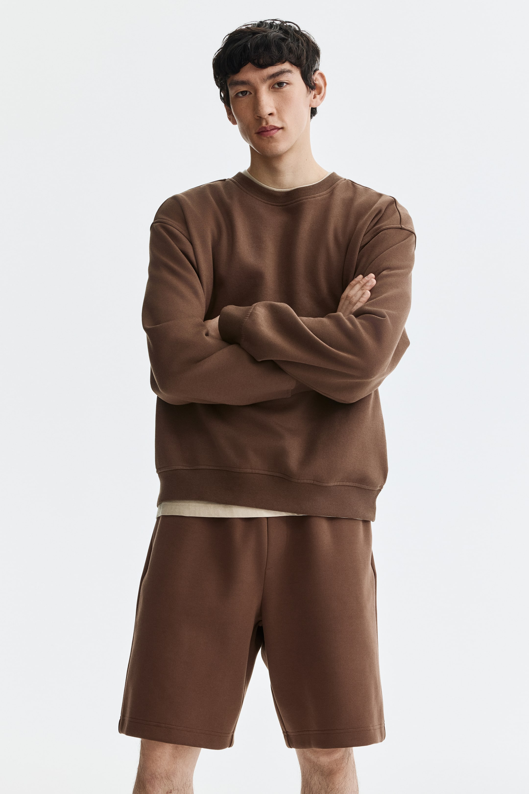 Größeres Bild ansehen: Ein braunes Sweatshirt mit Rundhalsausschnitt und passende braune Shorts werden von vorne gezeigt. Das locker sitzende Sweatshirt hat gerippte Bündchen und einen gerippten Saum, und darunter ist ein helles T-Shirt zu sehen. Die locker sitzenden Shorts haben Seitentaschen und reichen bis oberhalb des Knies.