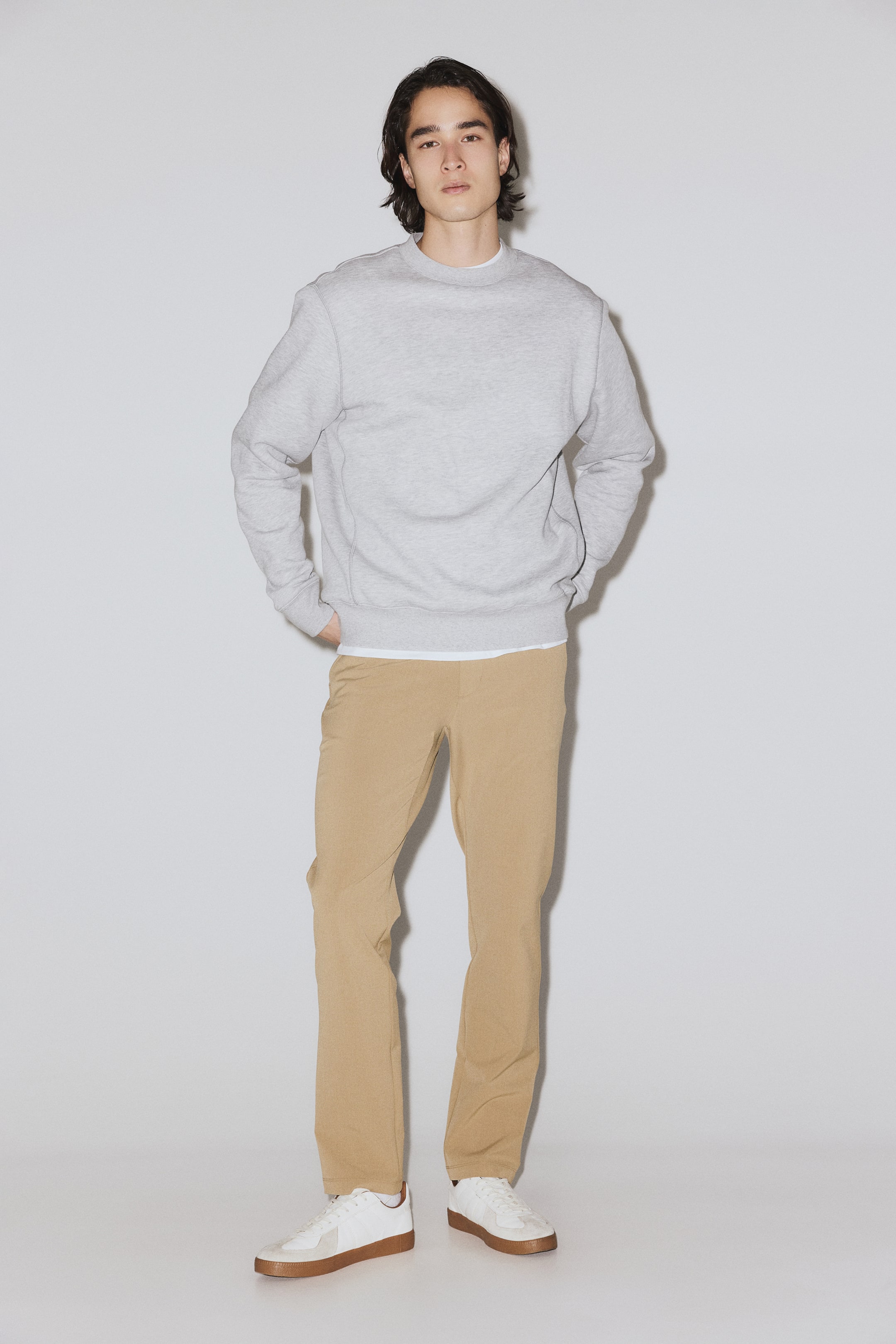 Ampliar la imagen: Una sudadera gris claro de cuello redondo presenta un ajuste holgado con puños acanalados y dobladillo acanalado; el cuello también es acanalado. Se lleva sobre una camiseta blanca, visible en el escote y en el dobladillo.