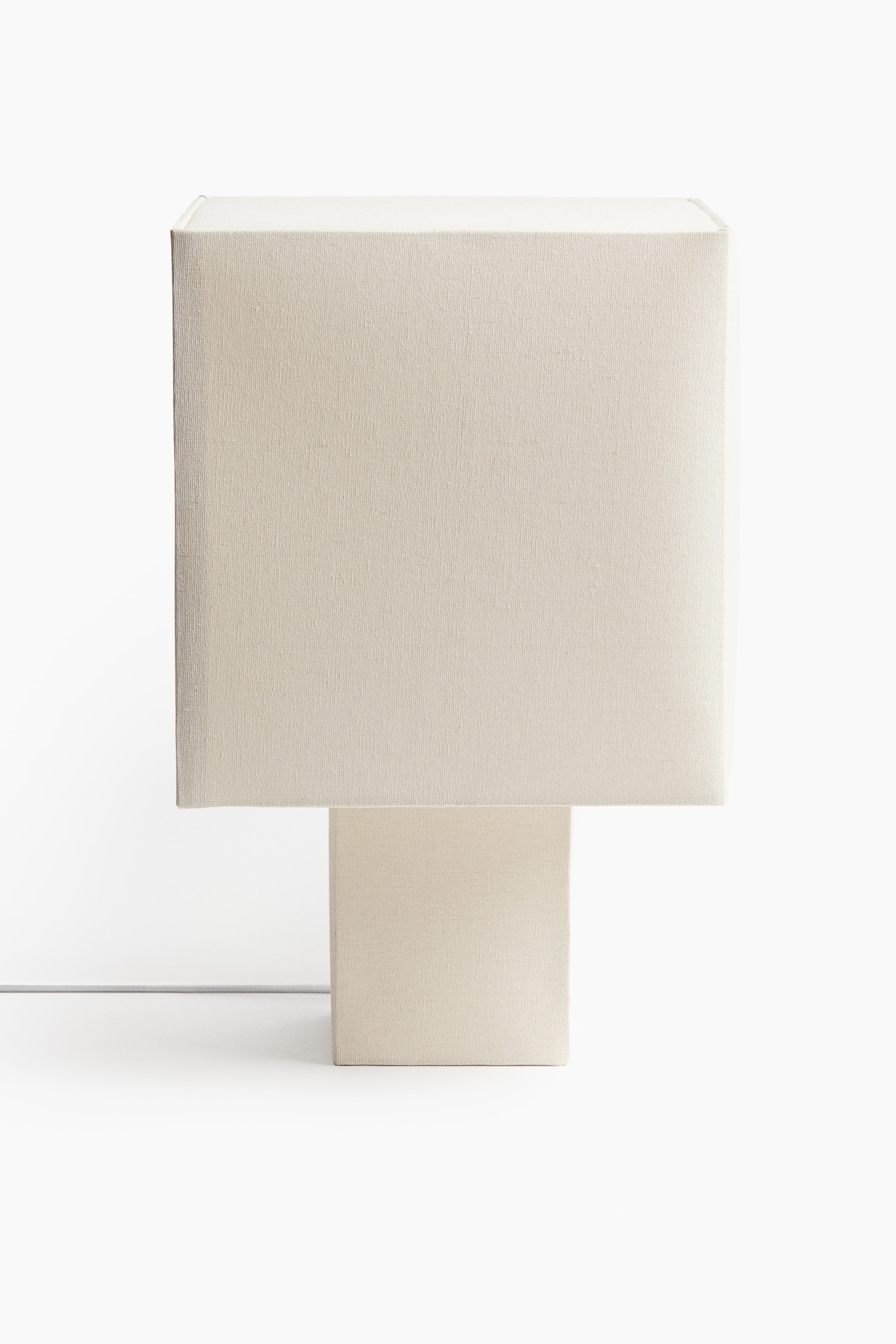 View larger image: A light beige table lamp features a large rectangular shade resting on a smaller rectangular base, both covered in textured fabric. A thin white cord extends from the lower left side of the base.