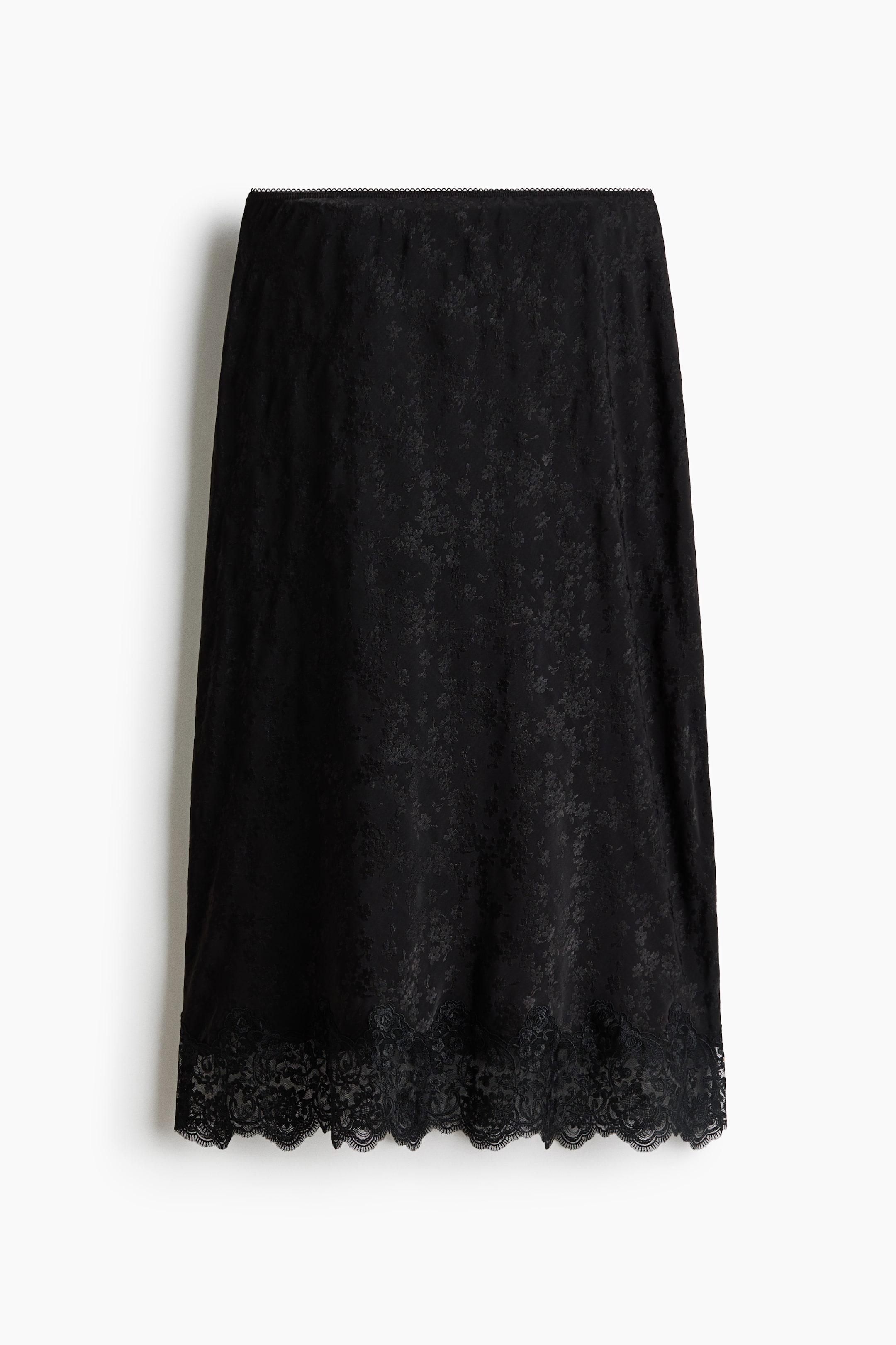 View larger image: A black slip skirt is shown from the front, featuring a subtle tonal floral jacquard pattern. The midi-length skirt is designed with a clean waistband and a wide, scalloped floral lace trim along the hem.