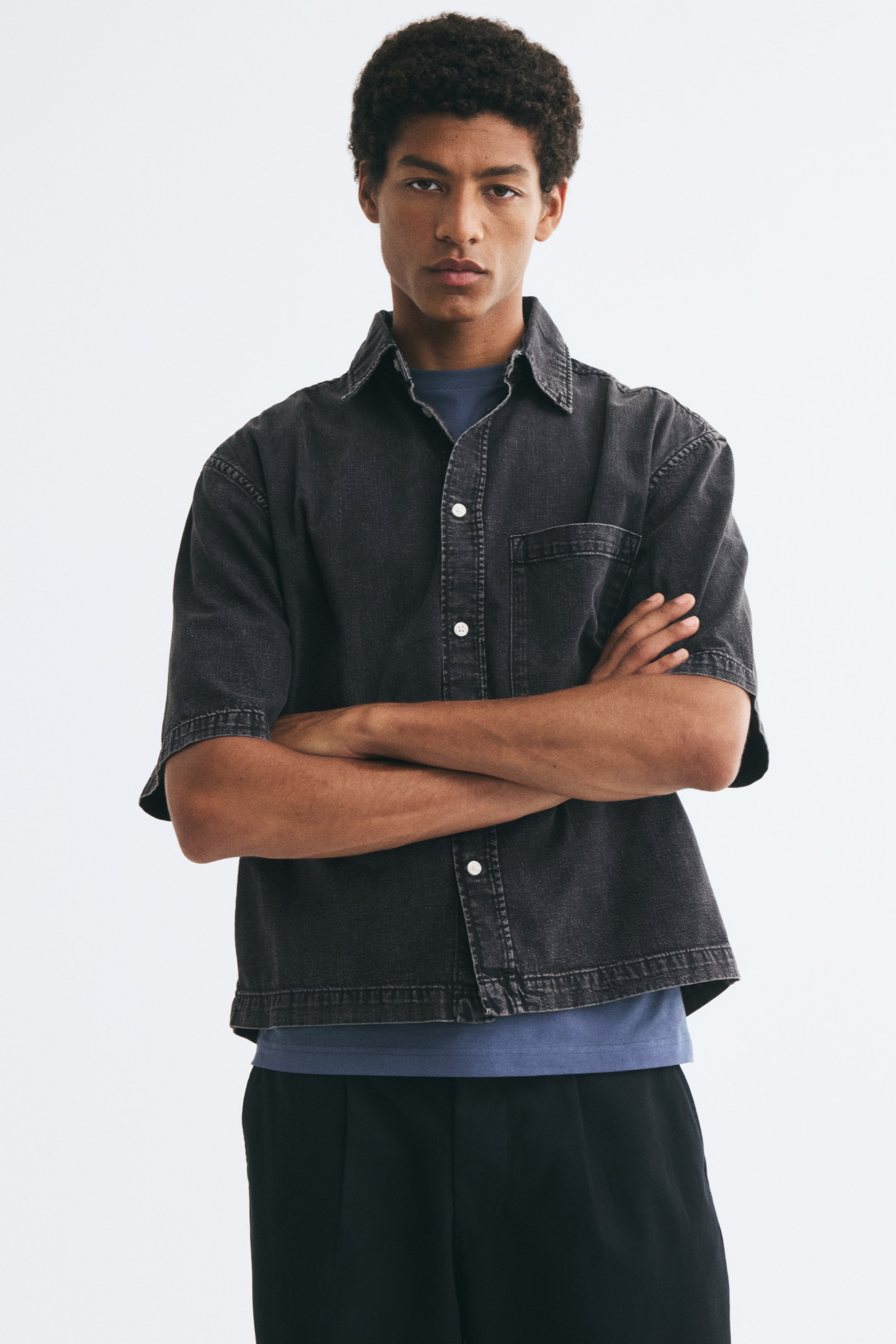 Xem ảnh lớn hơn: Người mẫu đang mặc áo sơ mi denim đen wash tay ngắn, đứng khoanh tay bên ngoài áo thun xanh và quần tây đen. Áo sơ mi dáng relaxed fit có cổ áo cổ điển, hàng cúc trắng phía trước, vai trễ và một túi ốp đơn trên ngực.