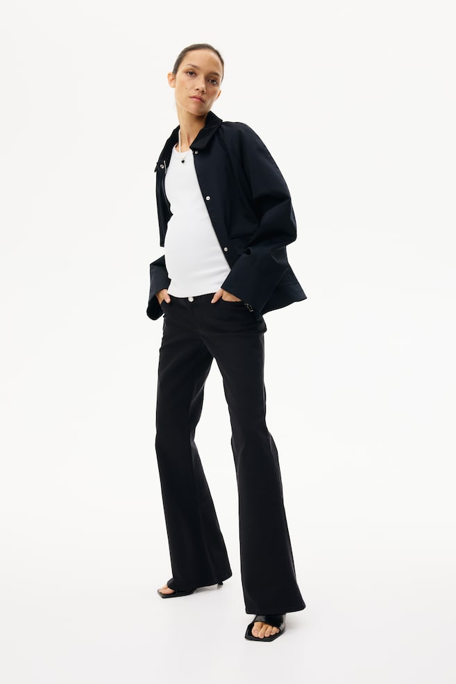 A black collared jacket is being worn open, revealing a white ribbed top underneath. Black wide-leg pants are extending to the floor, featuring a prominent flare from the knee. - 1
