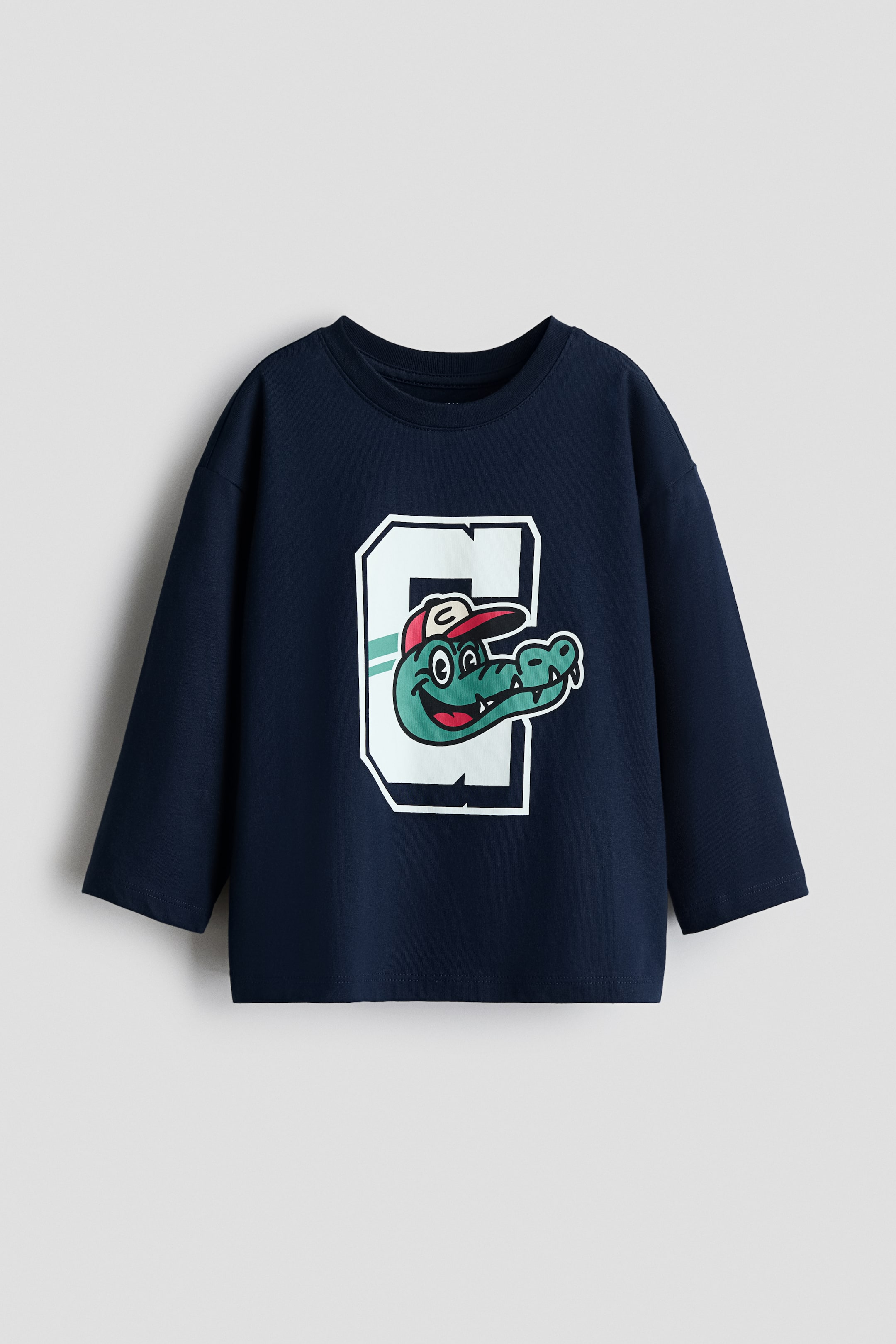 View larger image: A dark blue long-sleeved t-shirt featuring a crew neck and long sleeves. The front showcases a large graphic of a white outlined letter 'G' with a green stripe, and a cartoon green alligator head wearing a red and white baseball cap overlapping the 'G'.