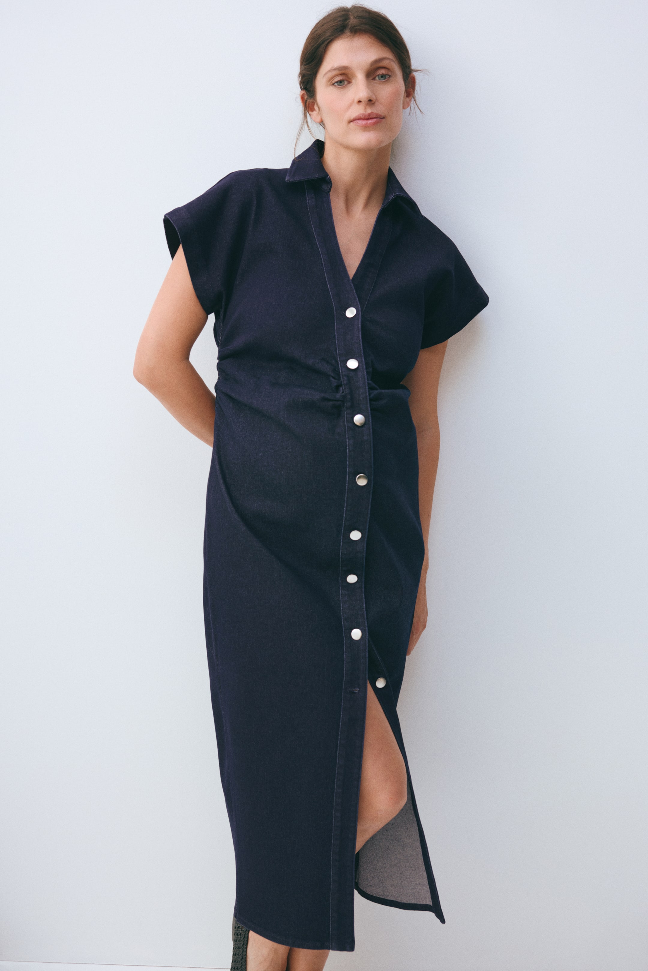 Nagyobb kép megtekintése: "Sötét mosású farmering ruha, amely hegyes gallérral, rövid ujjakkal és teljes gombos záródással rendelkezik világos színű gombokkal. A deréknál összeszedett, és elöl hasítékkal rendelkező, midi hosszúságú szoknyát mutat."