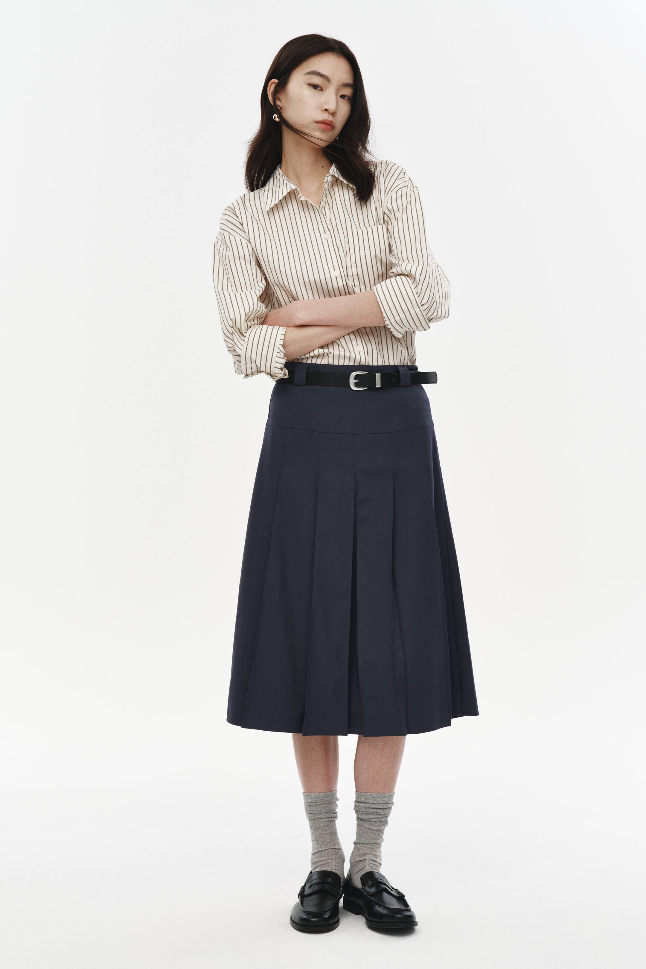 拡大画像を表示: クリーム色のシャツに細い縦のダークストライプが入ったデザインで、ポイントカラーとロールアップされた長袖が特徴です。ハイウエストのネイビーブルーのミディスカートにタックインされ、目立つプリーツがあり、黒いベルトでウエストが締められています。