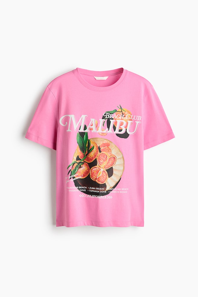 A pink cotton jersey t-shirt is laid flat, facing forward, featuring a round neckline and a straight-cut hem. The front is printed with "Malibu Beach Club" text, accompanied by illustrations of sliced and whole oranges or grapefruits and a list of California beach names. - 1