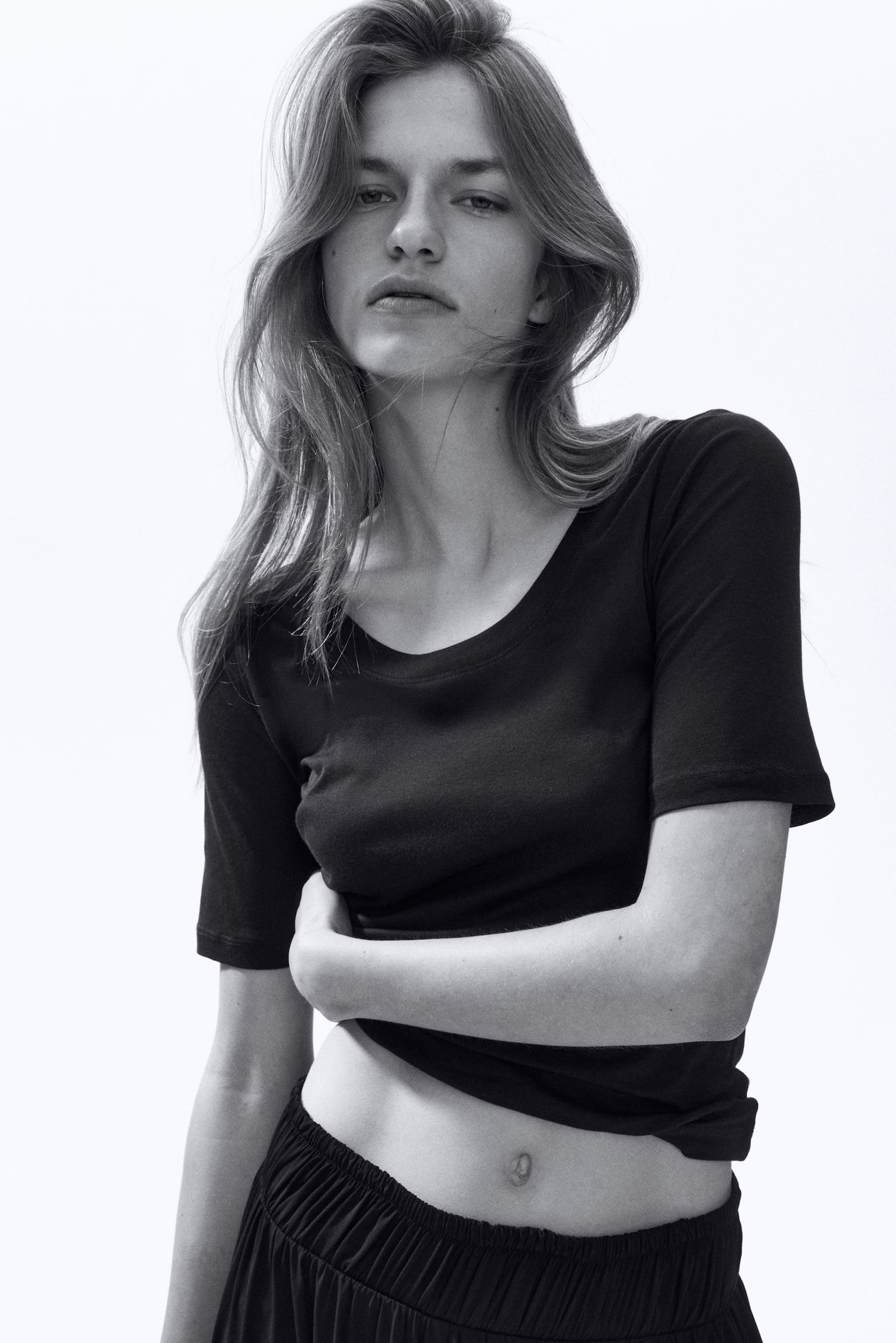 A black short-sleeved t-shirt featuring a scoop neck and a fitted silhouette, displayed from the front. It's styled with its hem pulled up, revealing black gathered trousers with an elasticated waist. - 1