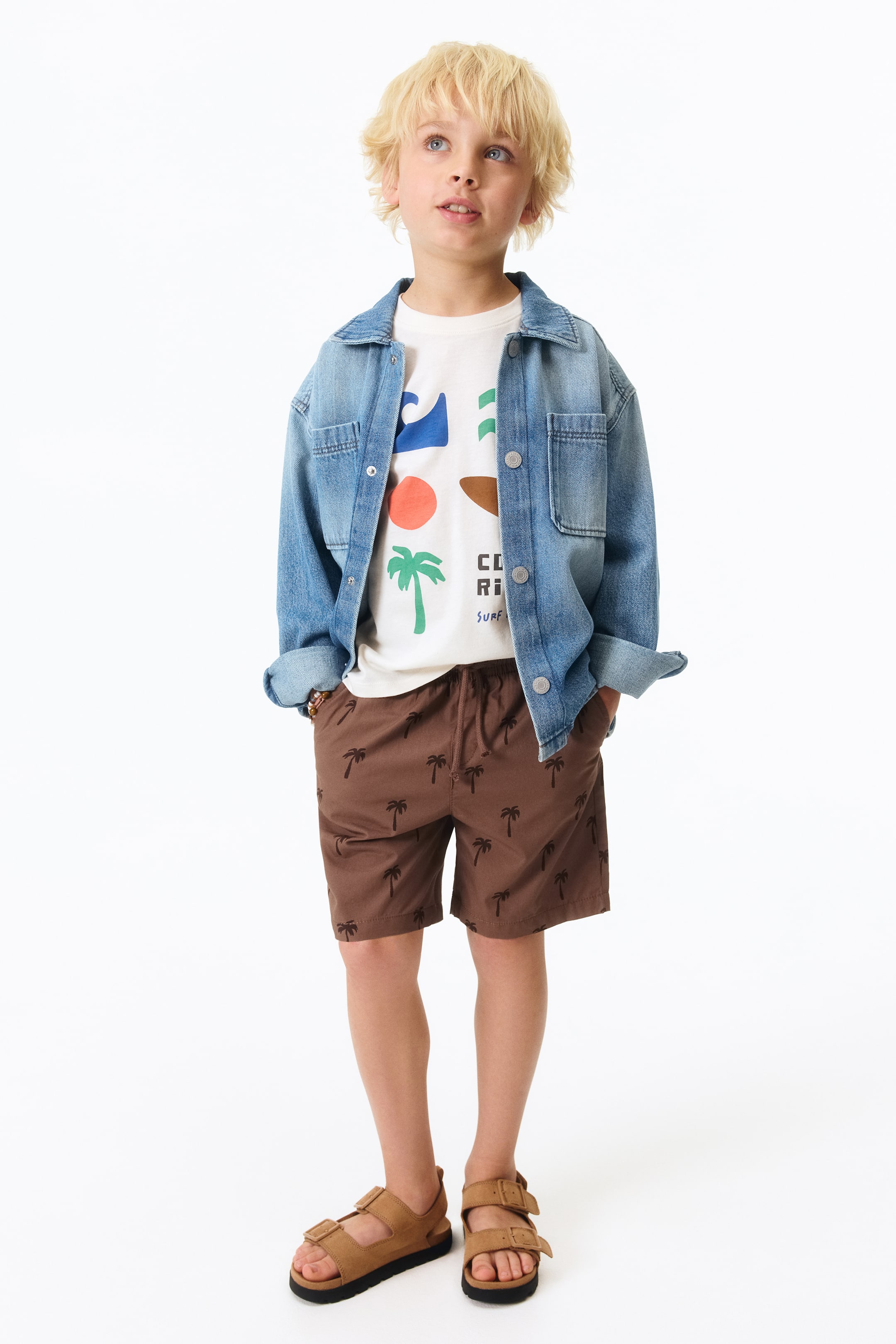 View larger image: A child's outfit features a light blue denim shirt jacket worn open over a white short-sleeved t-shirt with a colourful graphic print. Brown shorts with an elasticated drawstring waist and dark brown palm tree patterns are visible, completed by tan sandals with two buckled straps.