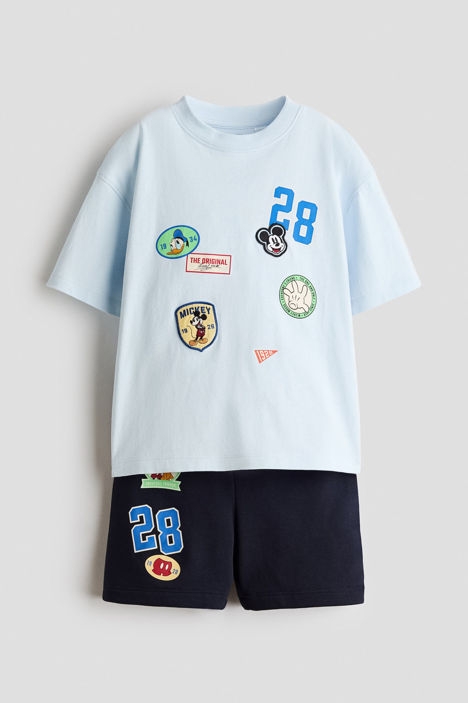 A two-piece clothing set is shown from the front, featuring a light blue, short-sleeved T-shirt with a ribbed crewneck, dropped shoulders, and various Disney-themed graphic designs on the front. The set includes coordinating navy blue sweat shorts with an elasticated waistband and matching graphics printed on the left leg.