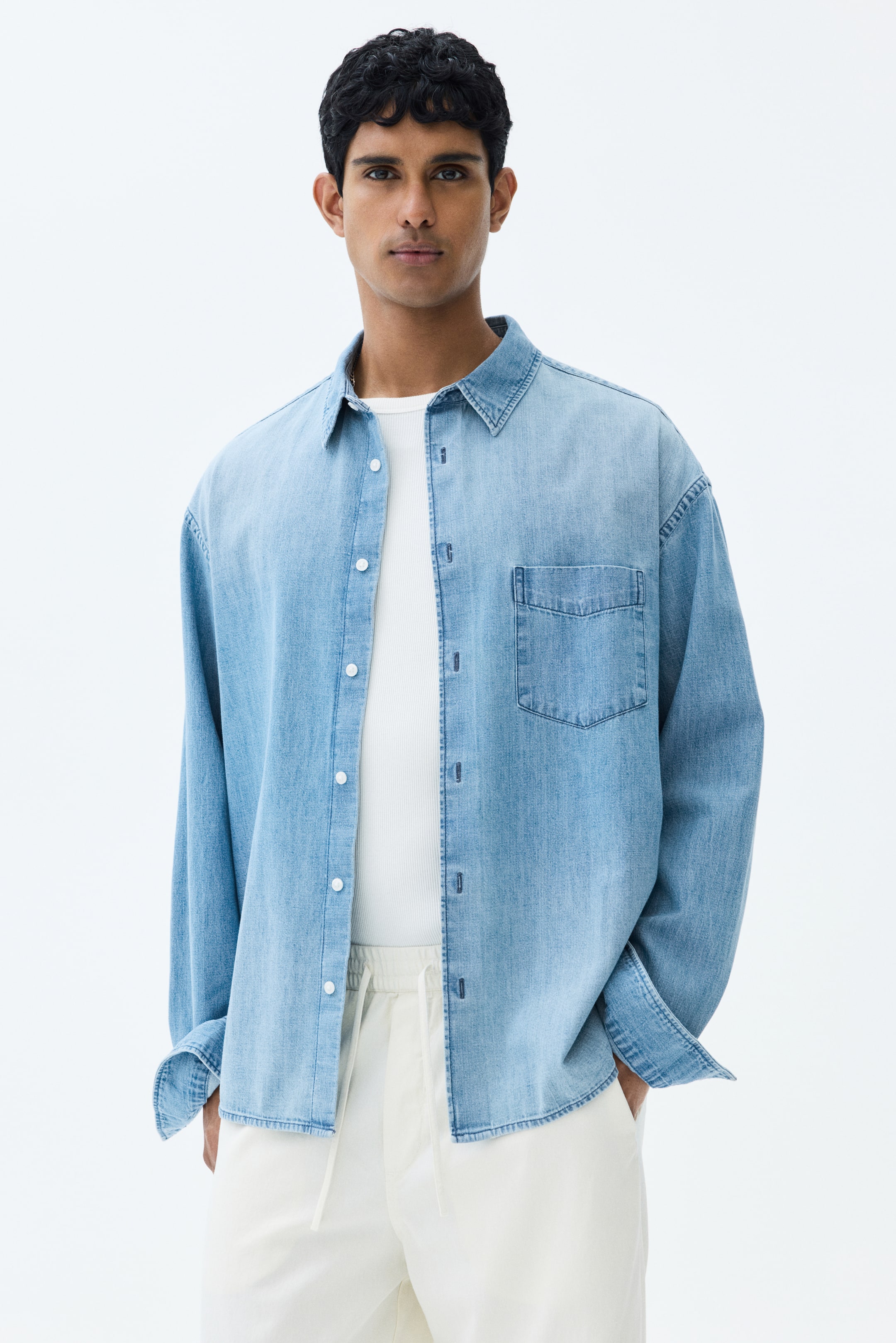 View larger image: The model is donning a light blue rigid cotton denim shirt with a relaxed fit, showcasing a collar, a French front, a patch chest pocket, and a subtly rounded hem. The shirt is worn open, revealing a white ribbed tank top beneath, and the long sleeves are rolled up at the cuffs.