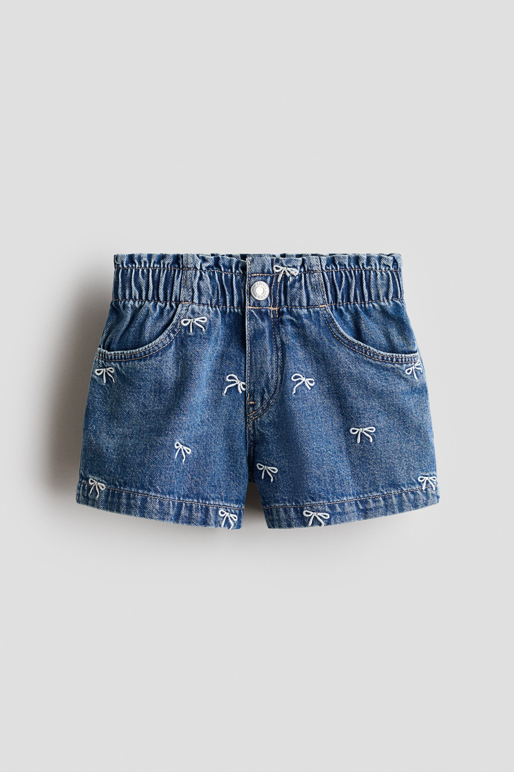 View larger image: Denim shorts are showcased from the front, featuring a blue denim fabric adorned with scattered white embroidered bows. They are crafted with an elasticated, gathered waistband, a single silver-toned button, and two curved front pockets.